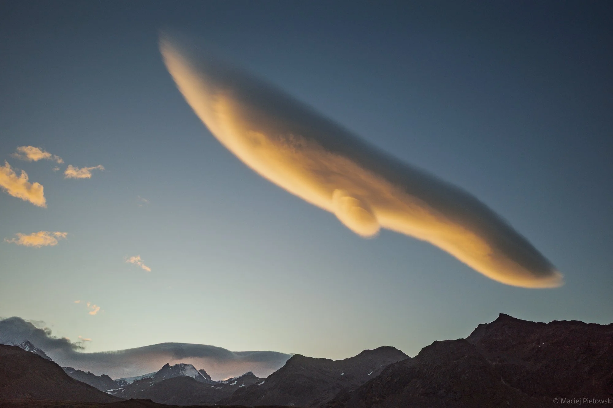 A mountain landscape at sunset with tall, rugged mountains under a sky with a large, curved cloud illuminated with yellow-orange light.