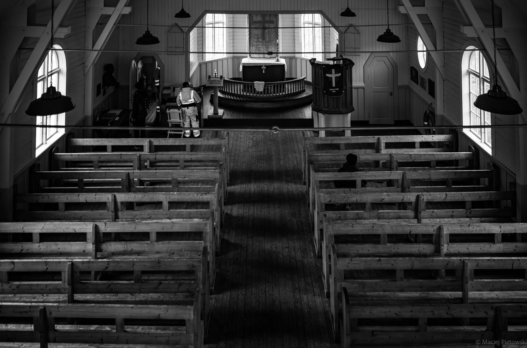 Interior of a church with empty wooden pews and a small balcony with a pulpit. People are standing near the altar, and a kid is sitting on a pew. Large windows provide natural light, and pendant lights hang from the ceiling.