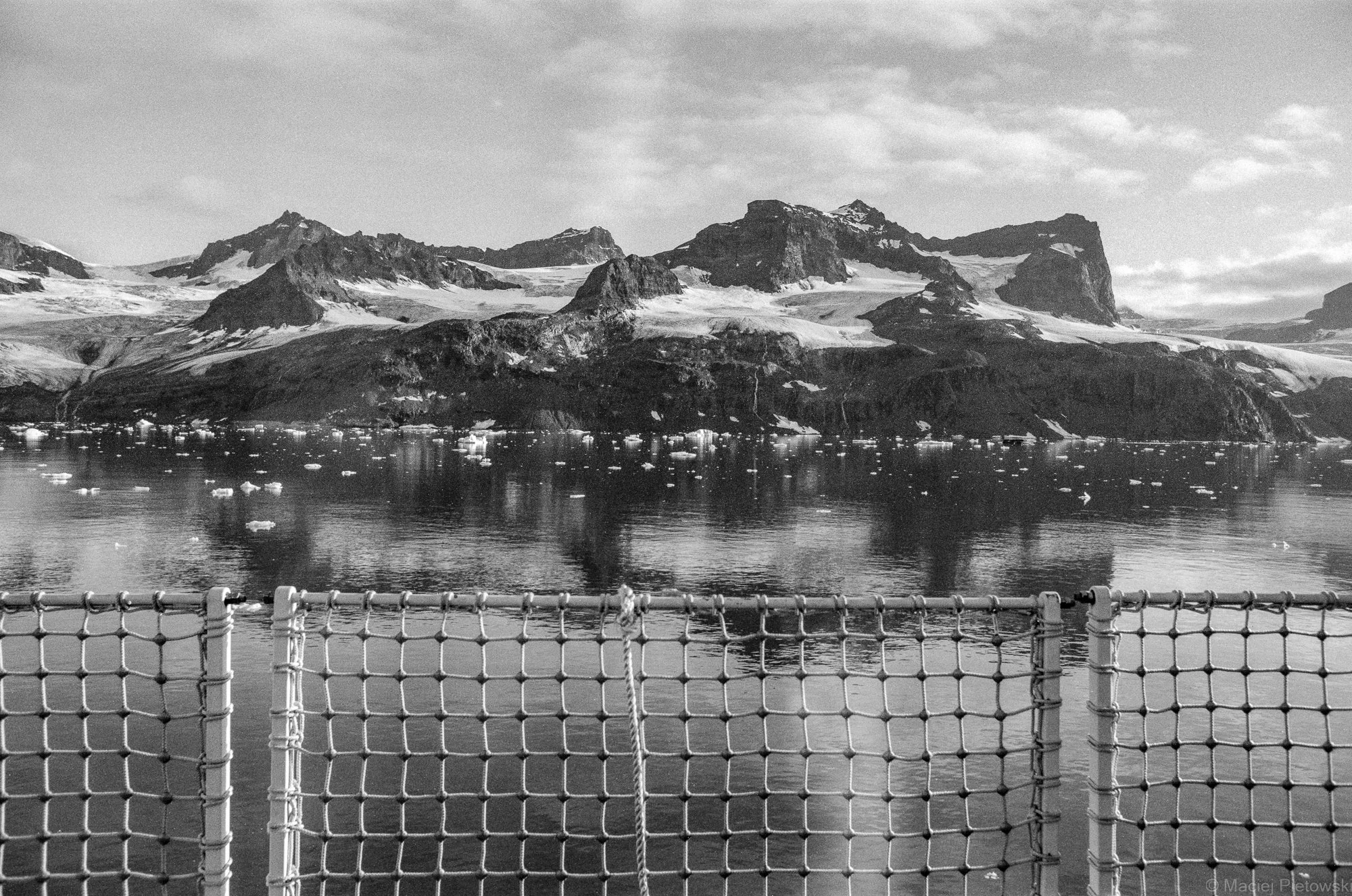 Black and white photo of a mountain landscape with glaciers and ice floating in the water, taken from a boat with a safety net in the foreground.