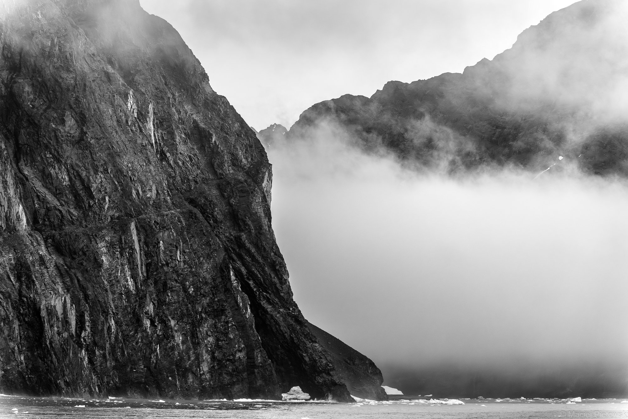 Black and white photo of rugged mountain cliffs rising from the shoreline with mist or fog surrounding them and a small, white boat near the base of the cliffs.