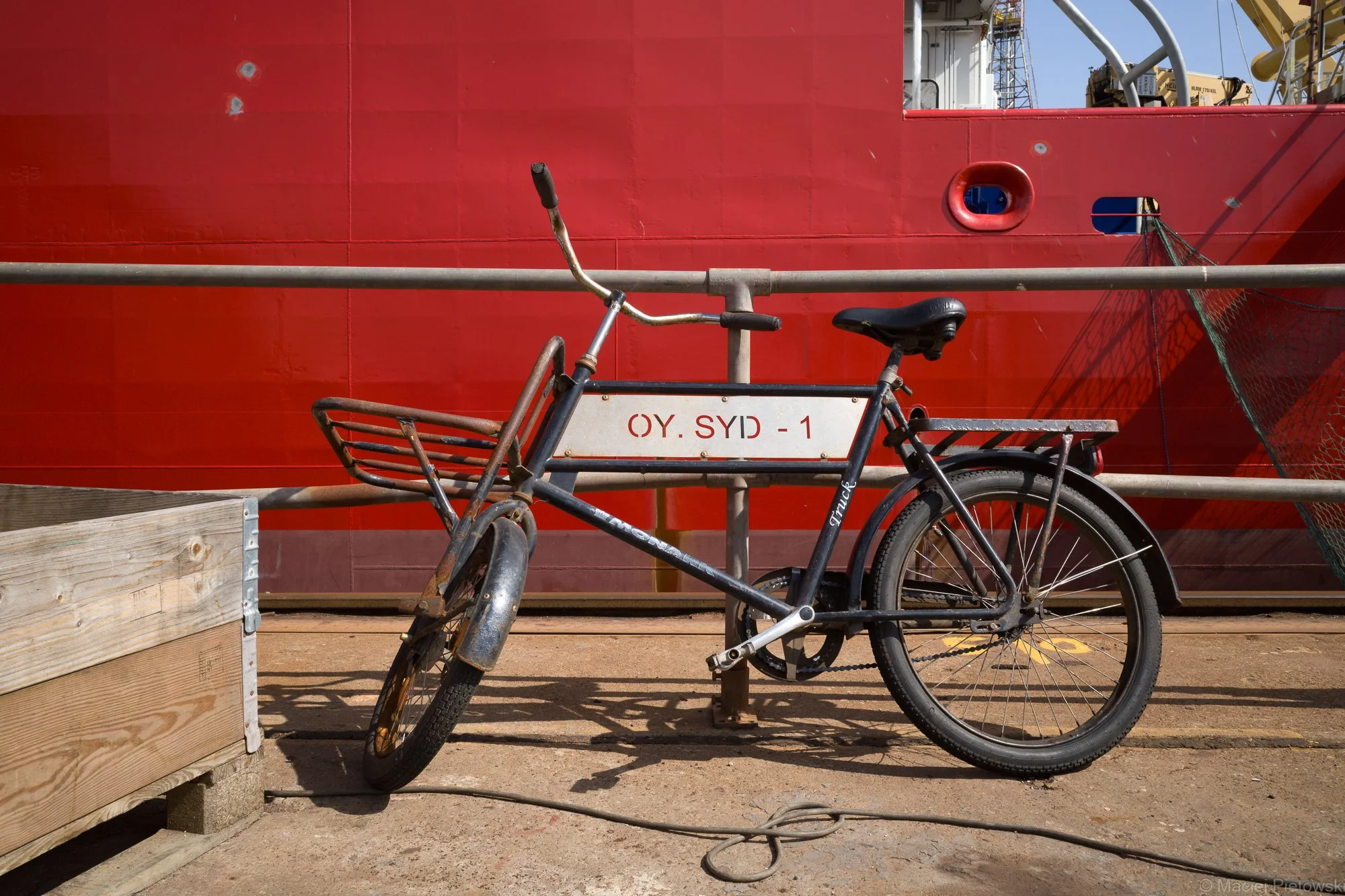 Old black bicycle parked in front of a large red vessel with a cutout porthole, and construction equipment visible in the background.