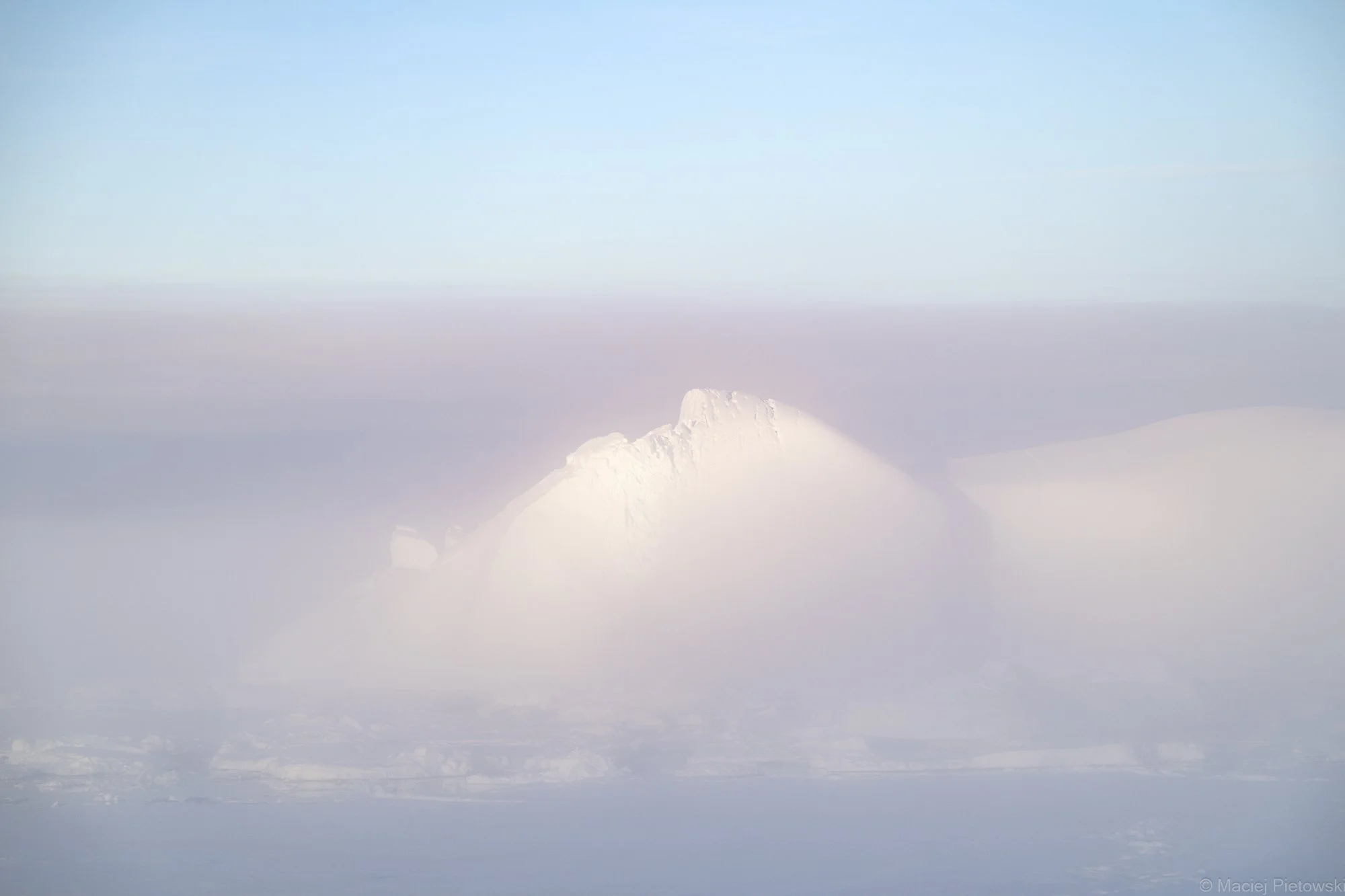 Snow-covered mountain peak surrounded by fog or clouds under a clear blue sky.