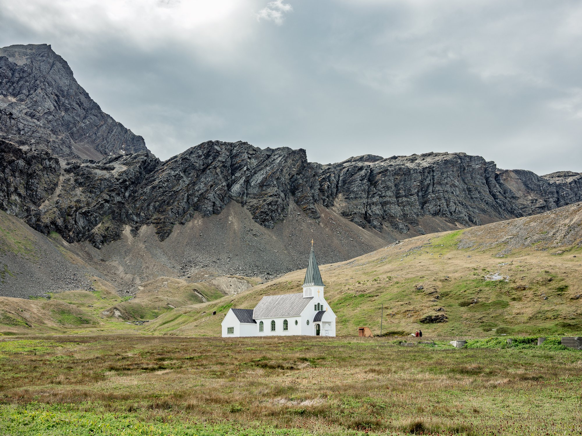 A white church with a pointed steeple is situated in a green field in front of tall, rugged mountains under a cloudy sky.