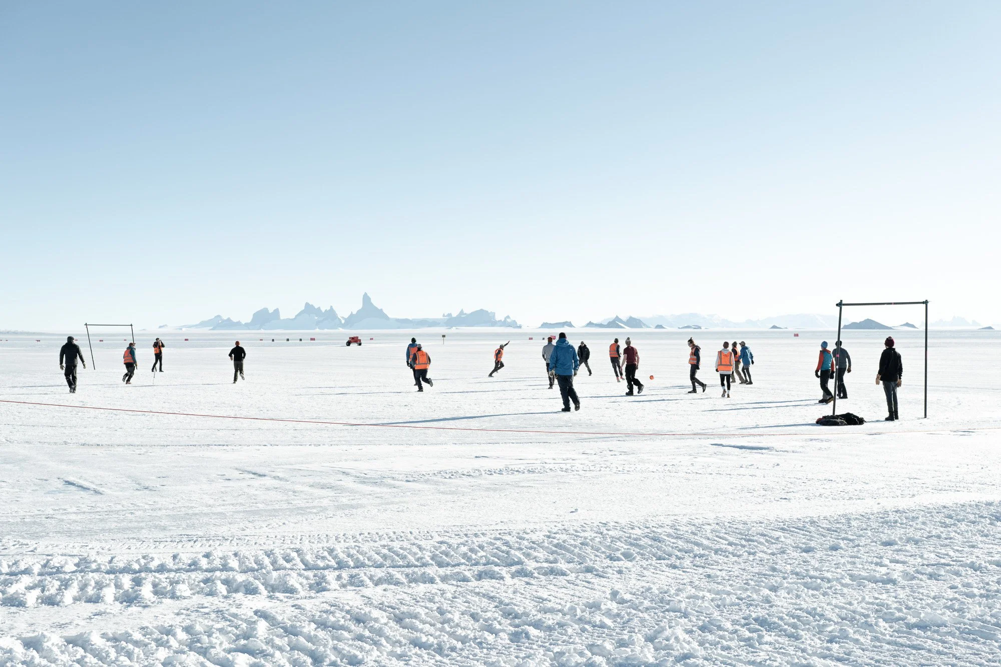 People playing soccer on a snowy field with mountains in the background under a clear blue sky.