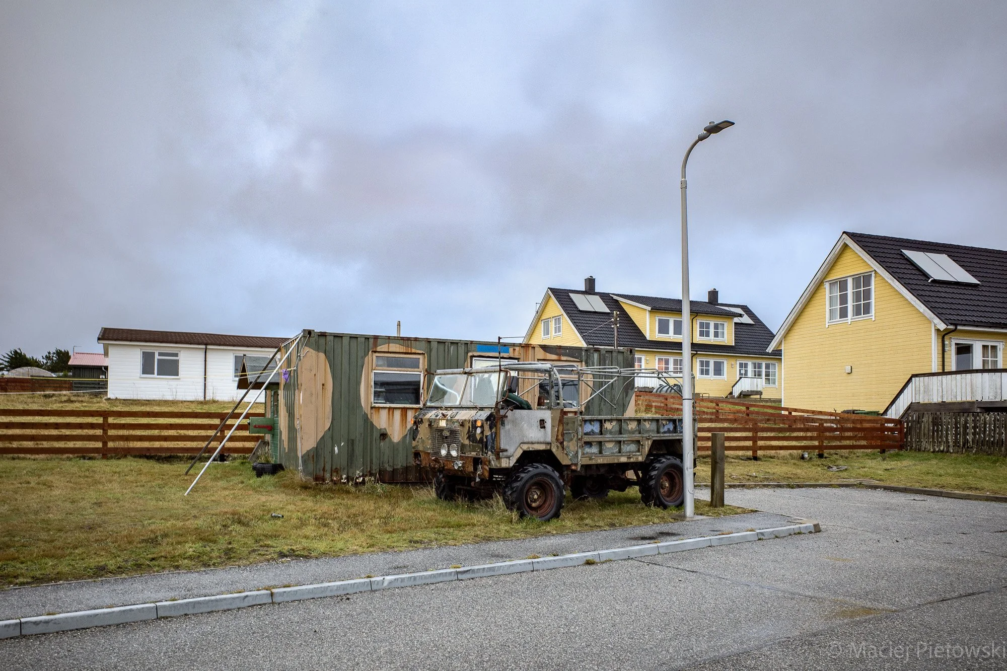 A rusty military-style jeep parked on a grassy area next to a sidewalk in front of yellow houses with black roofs, during overcast weather.