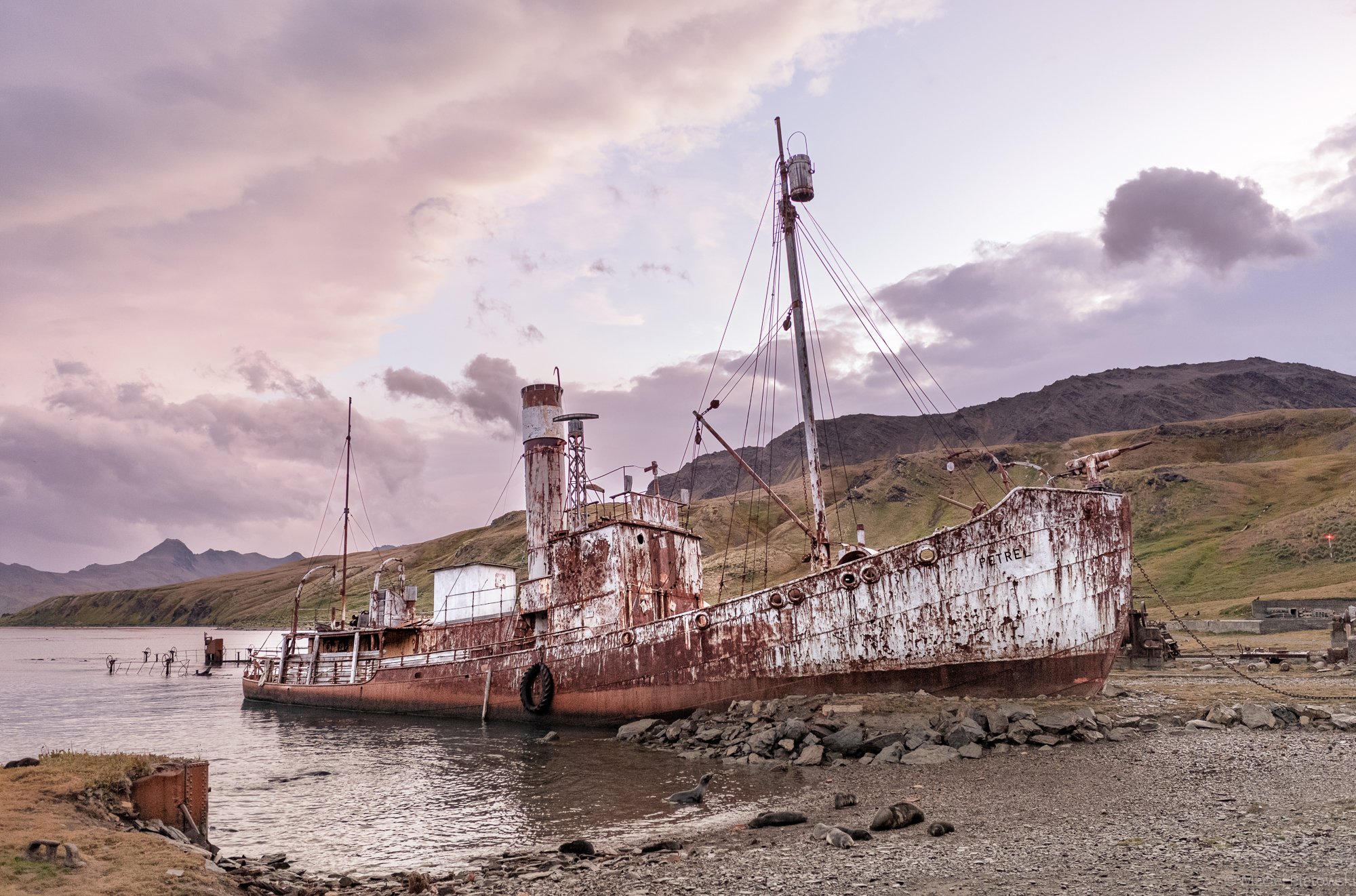 An old, rusted ship named PETREL beached on the rocky shoreline of a lake, with green hillside and mountains in the background under cloudy sky.