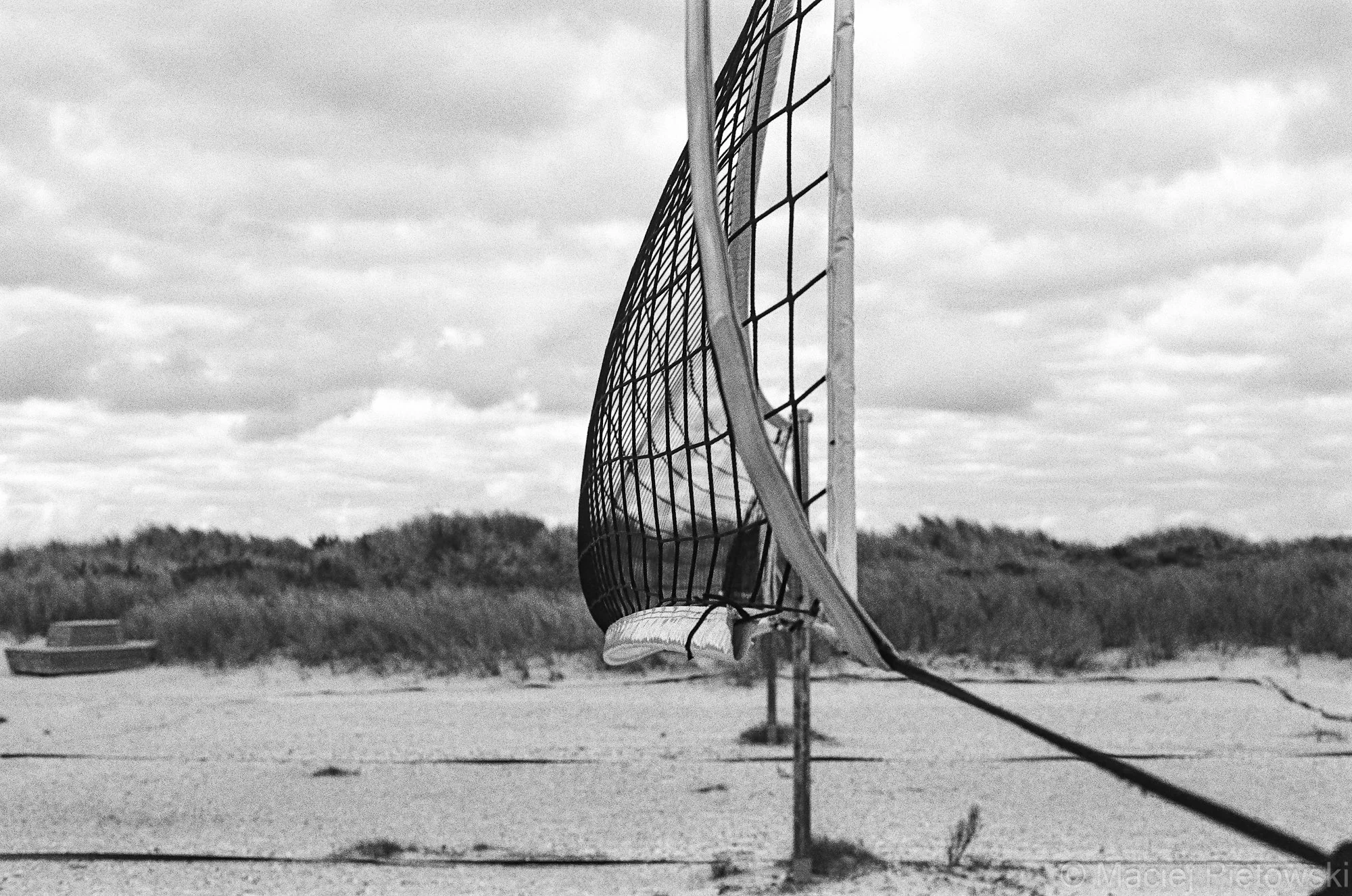 An outdoor volleyball net and pole on a sandy beach with grass and bushes in the background, under a cloudy sky.