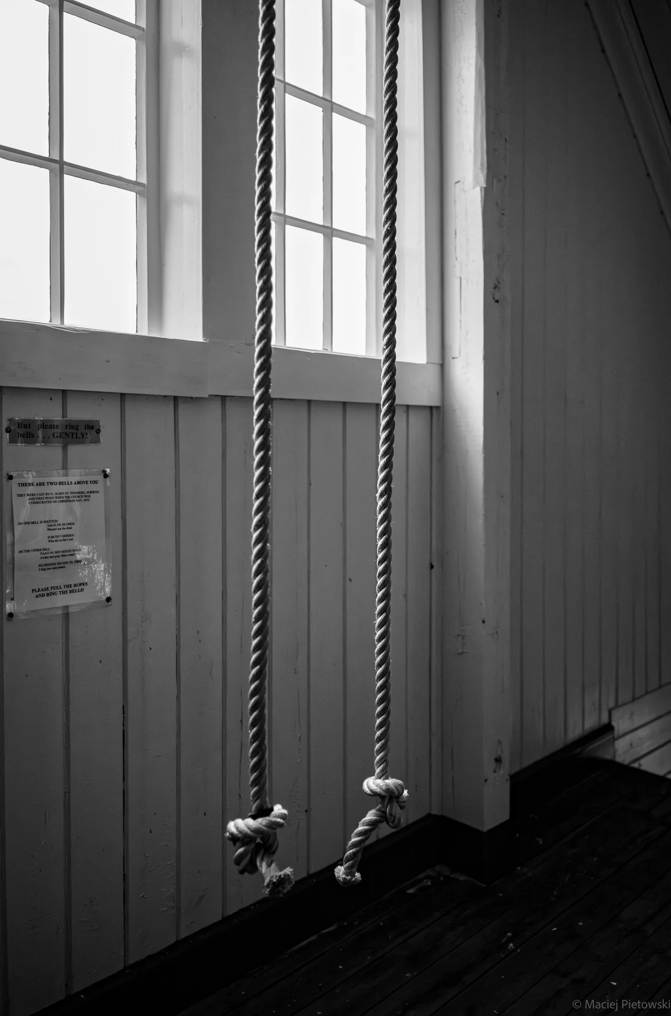 Black and white photo of a swing with two ropes hanging from a ceiling, in front of a window with multiple panes and a wall with a sign.