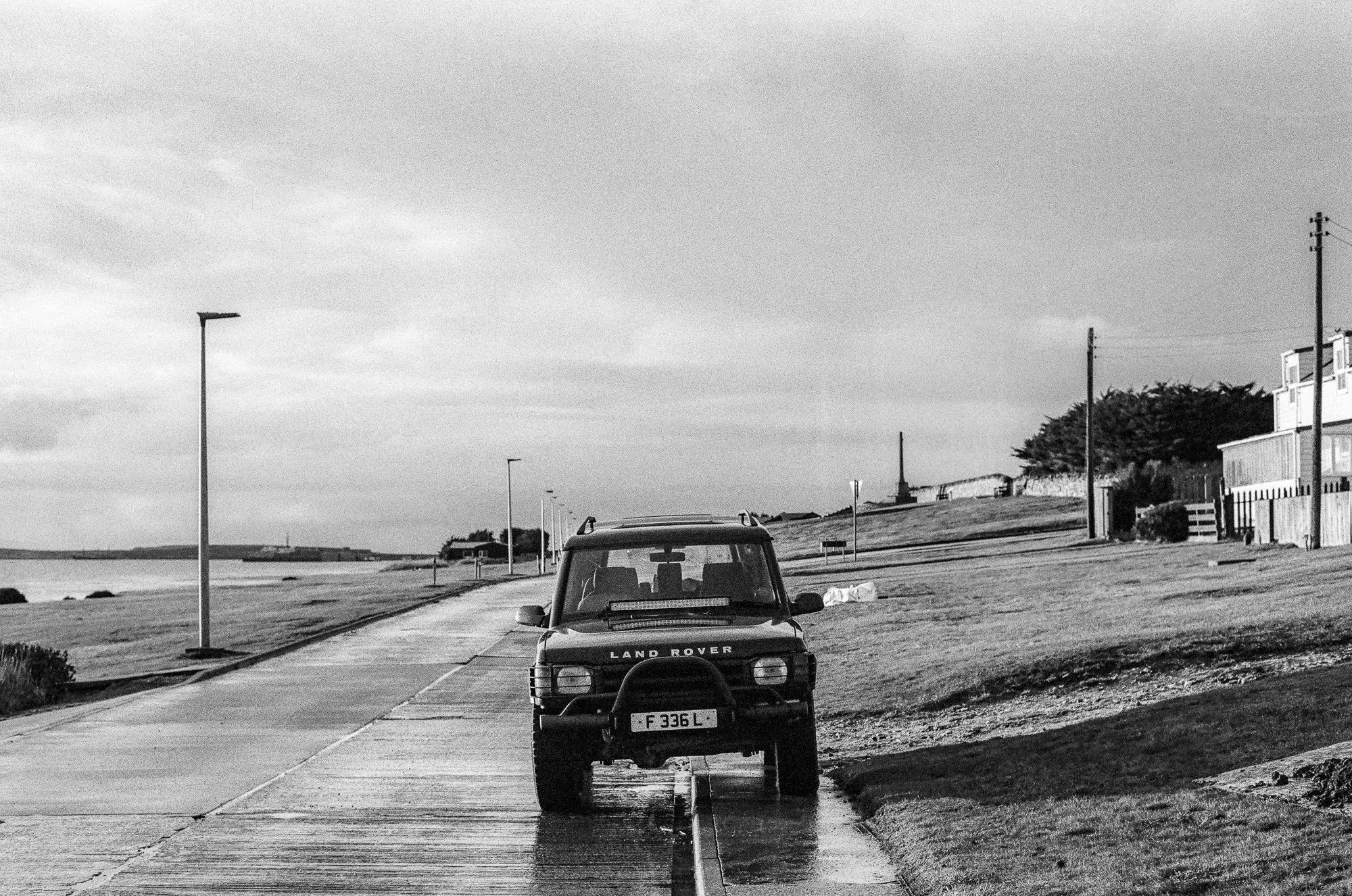 A black and white photo of a parking area near a coastline with a car in the foreground, houses, trees, and a lighthouse in the background. There are streetlights along the road and muddy patches on the ground.