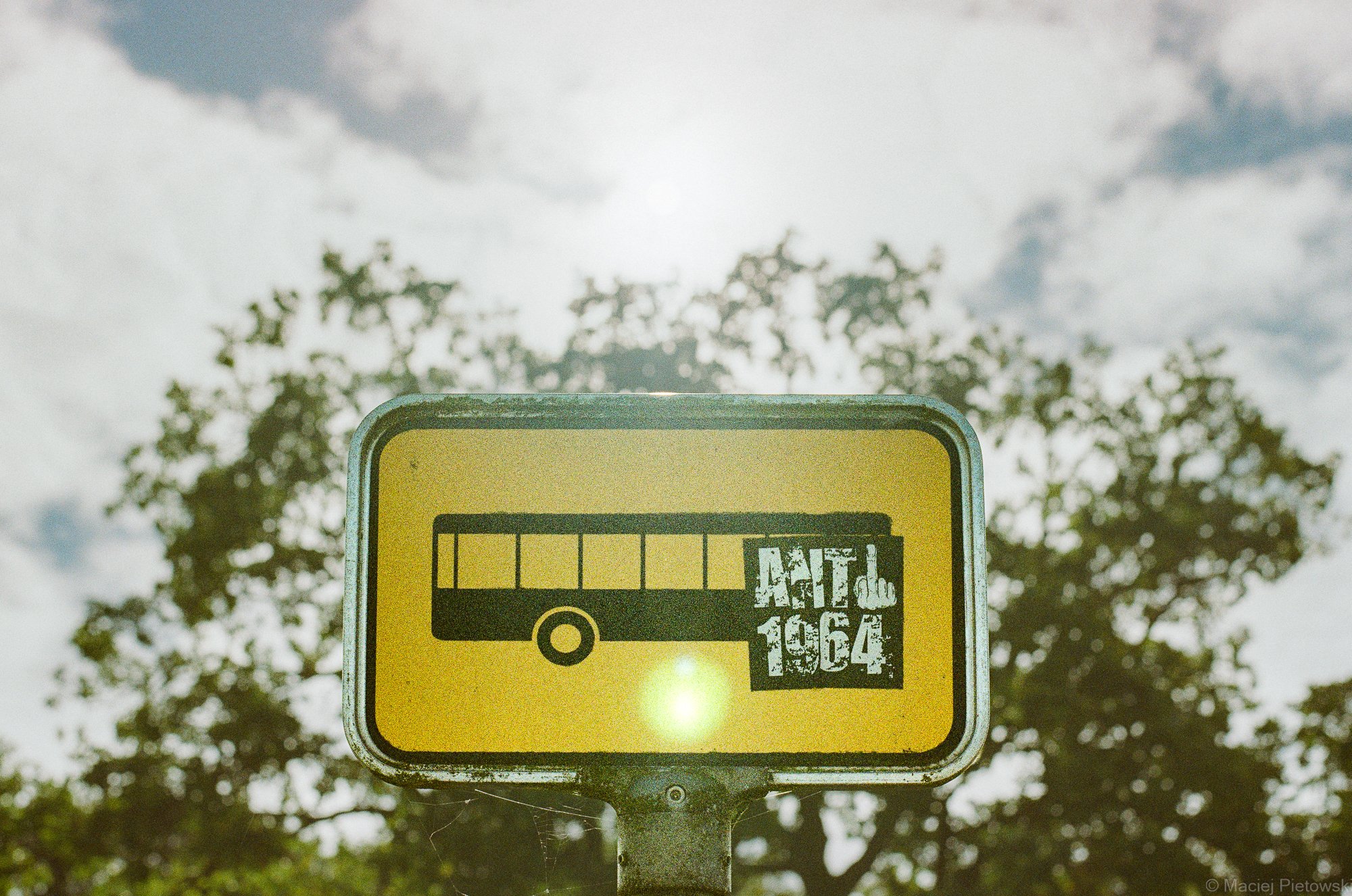 A yellow bus stop sign with a bus icon and text "ATU 1964," set against a background of trees and cloudy sky.