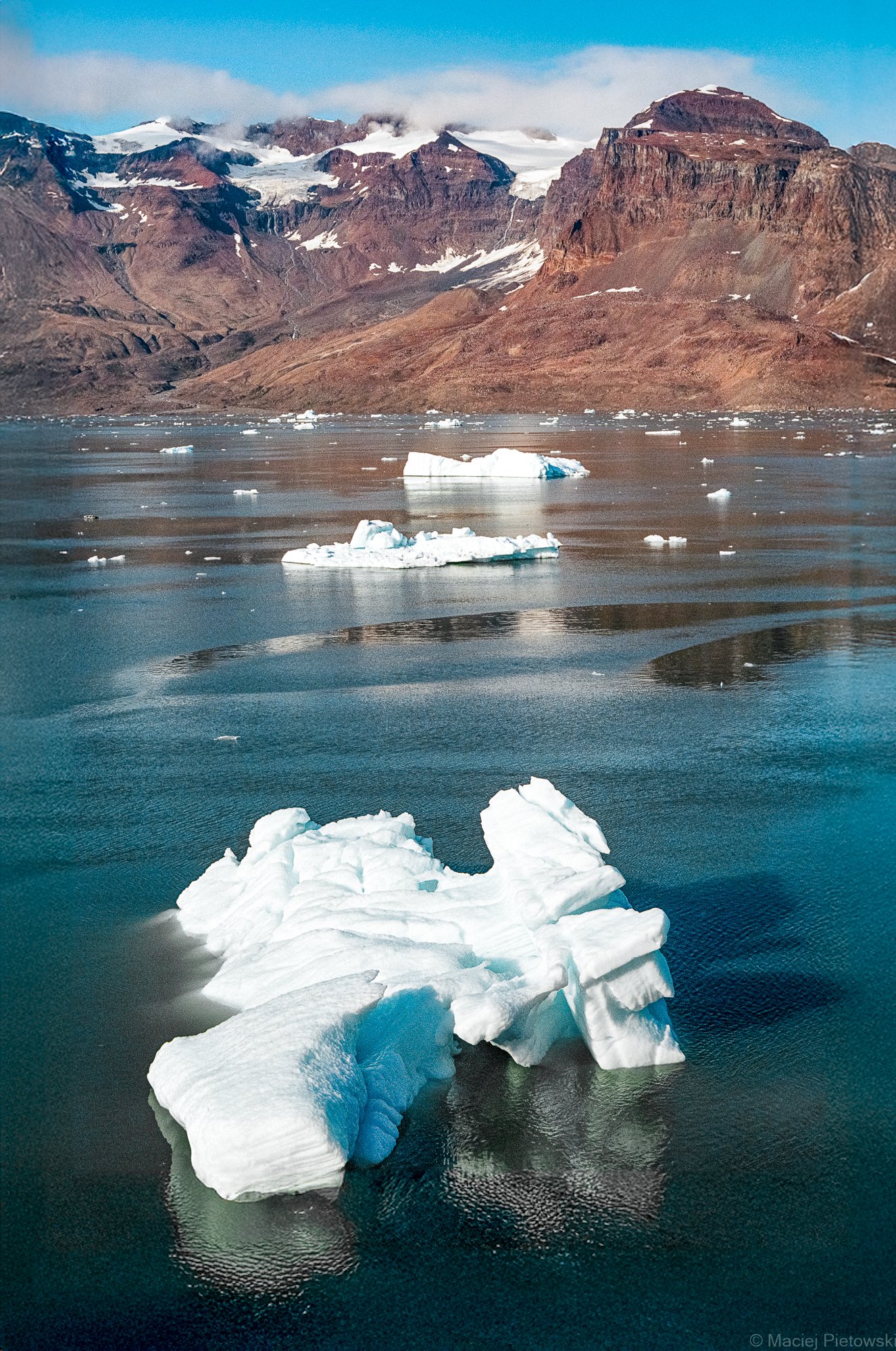 A large iceberg floating in cold, dark water near a mountain landscape with snow-capped peaks and brown rocky slopes under a partly cloudy sky.