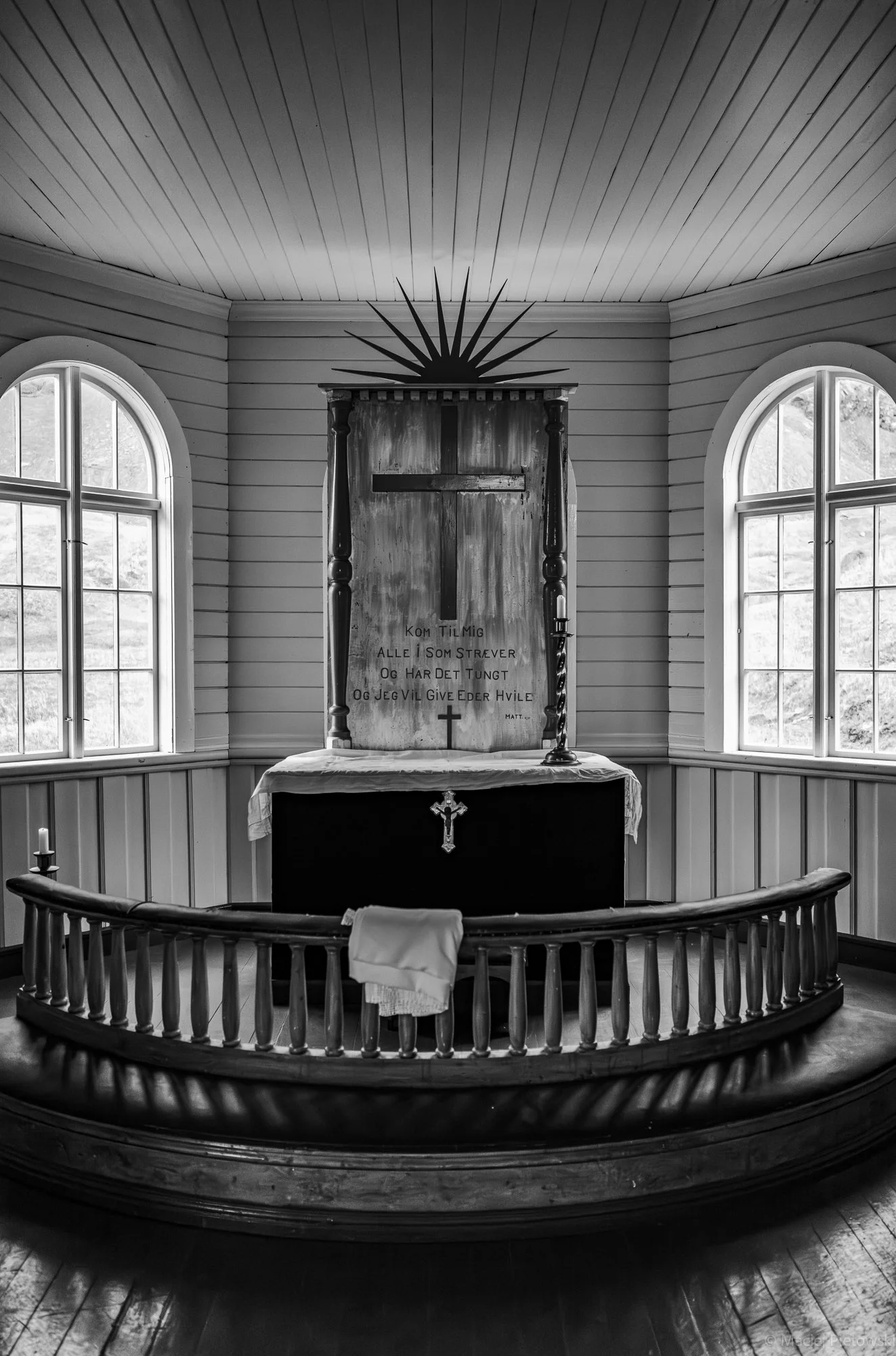 Black and white photo of an altar inside a church with a cross, candles, and a quote in a wooden frame, surrounded by two arched windows, a wooden railing, and wood-paneled walls and ceiling.
