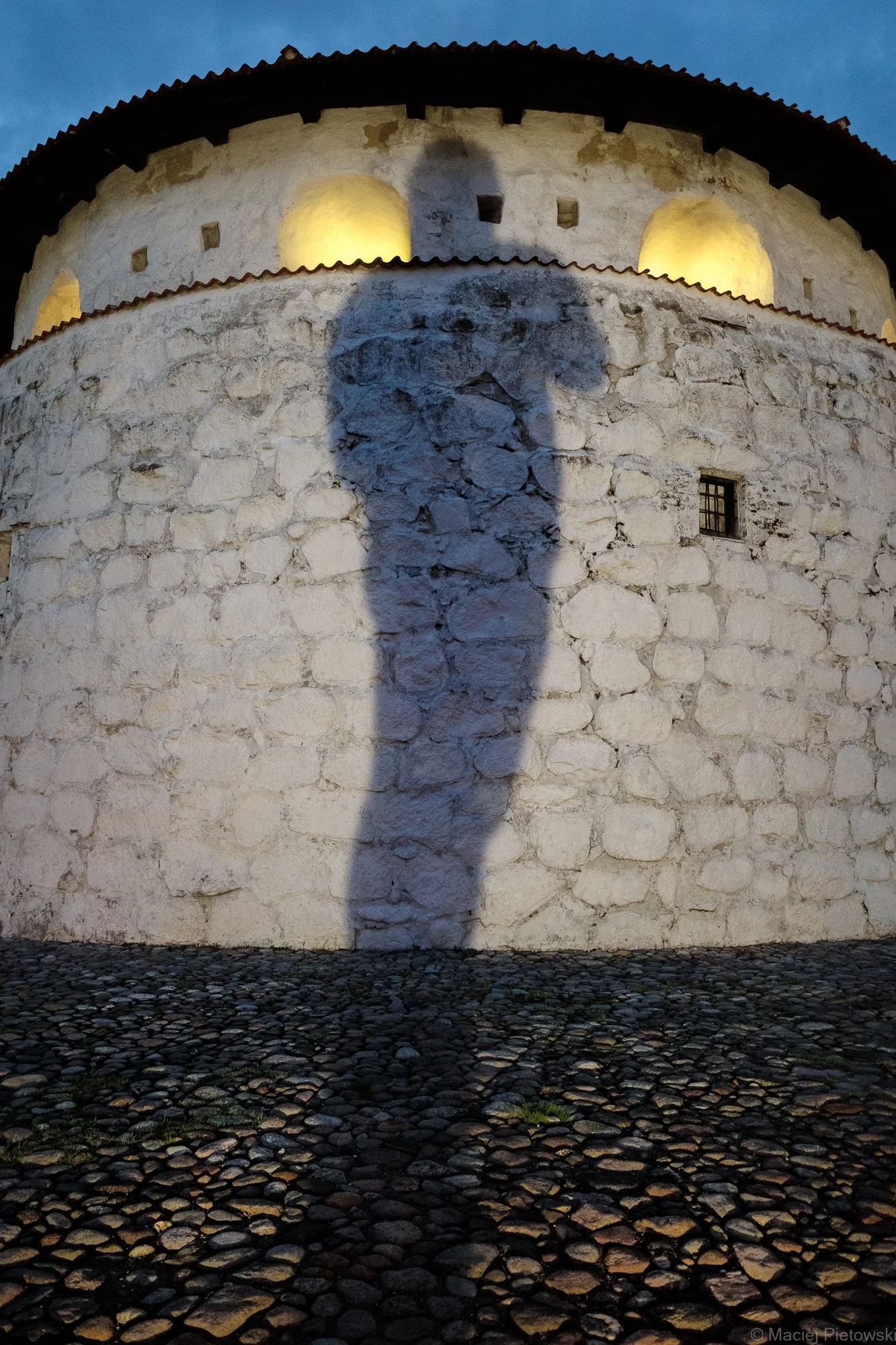 Shadow of a person cast on an ancient stone structure illuminated at dusk, with cobblestone ground in the foreground.