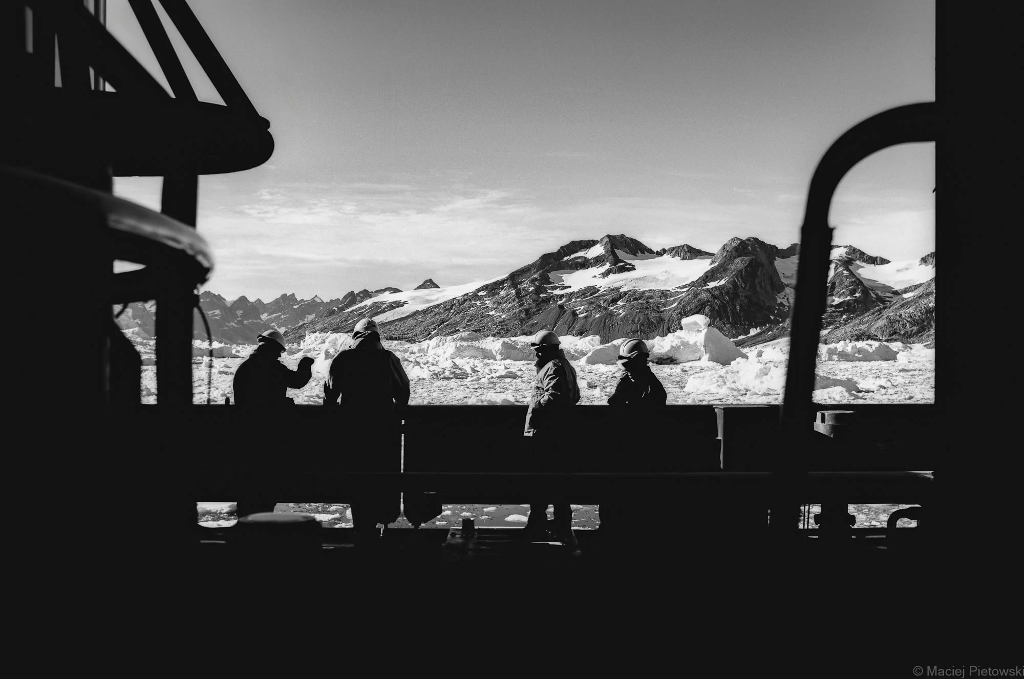 Black and white photograph of five people in winter gear, viewing a snowy mountain landscape through a window or opening, with visible snow, mountain peaks, and sky in the background.