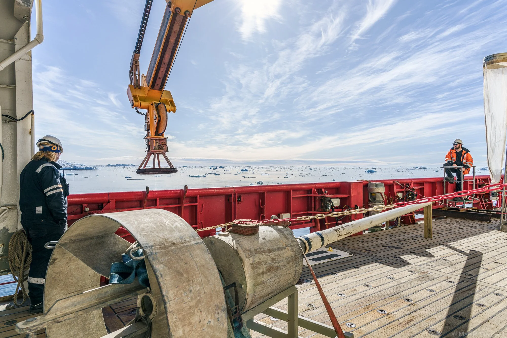 Two workers in safety gear operate equipment on a ship in icy waters under a partly cloudy sky.