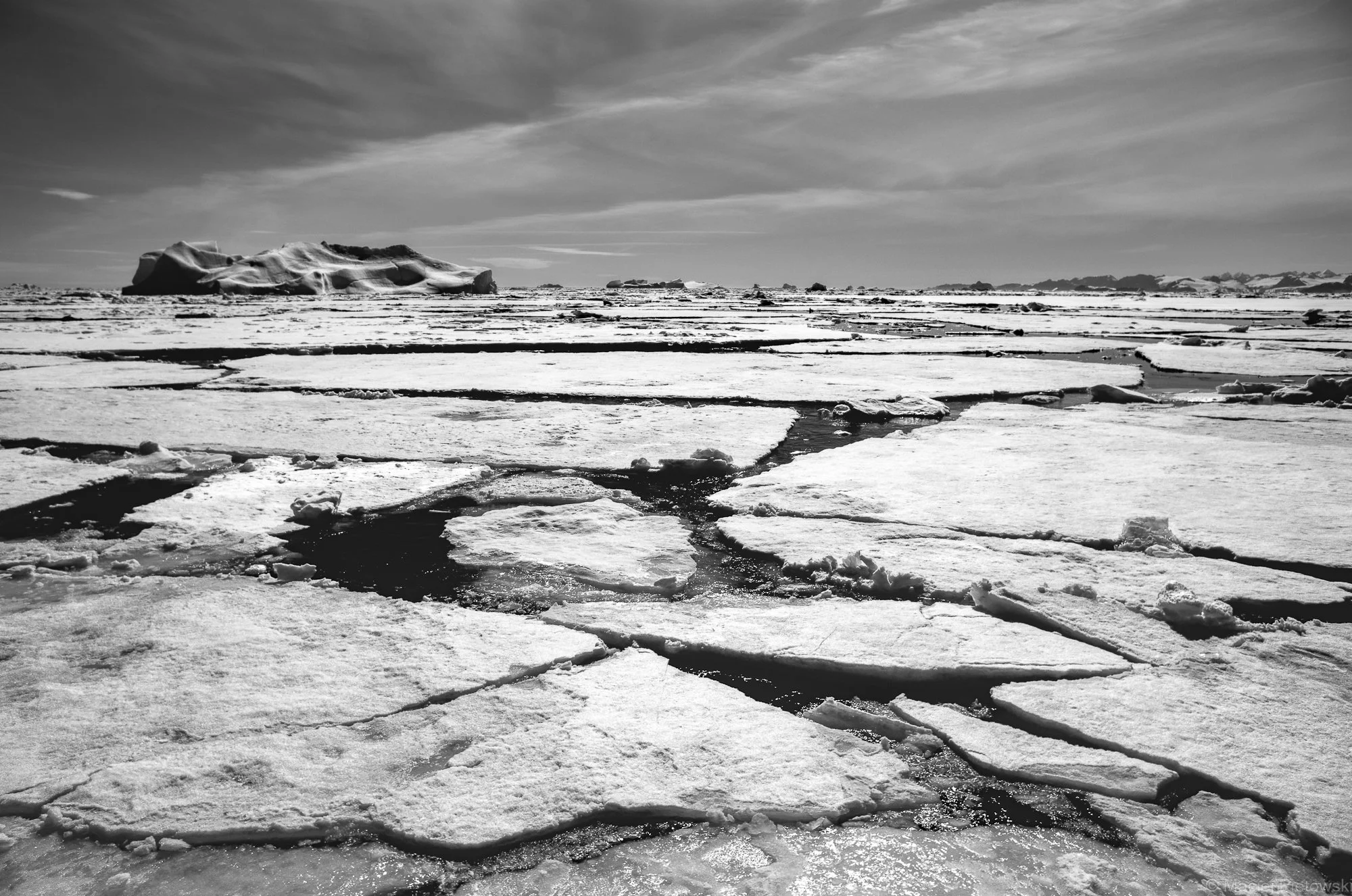 A black and white photograph depicting a vast icy landscape with large floating icebergs and cracked, frozen water surface extending to the horizon. Low clouds stretch across the sky, casting a subdued light.