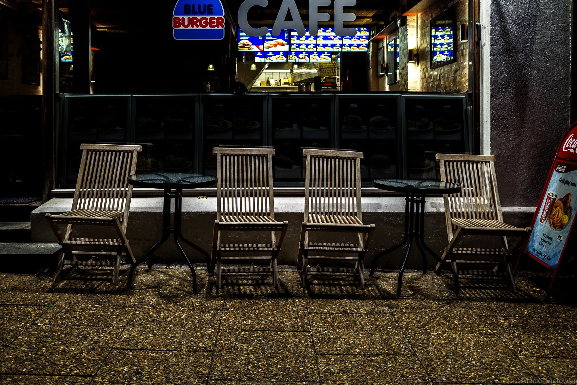 Empty outdoor seating area with wooden chairs and small round tables in front of a fast food restaurant with a bright menu displayed on digital screens.