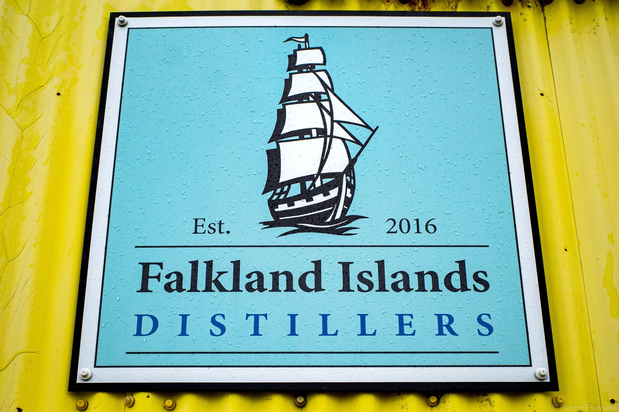 Sign for Falkland Islands Distillers featuring a black outline drawing of a sailing ship on a blue background, with yellow sides and water droplets on the surface.