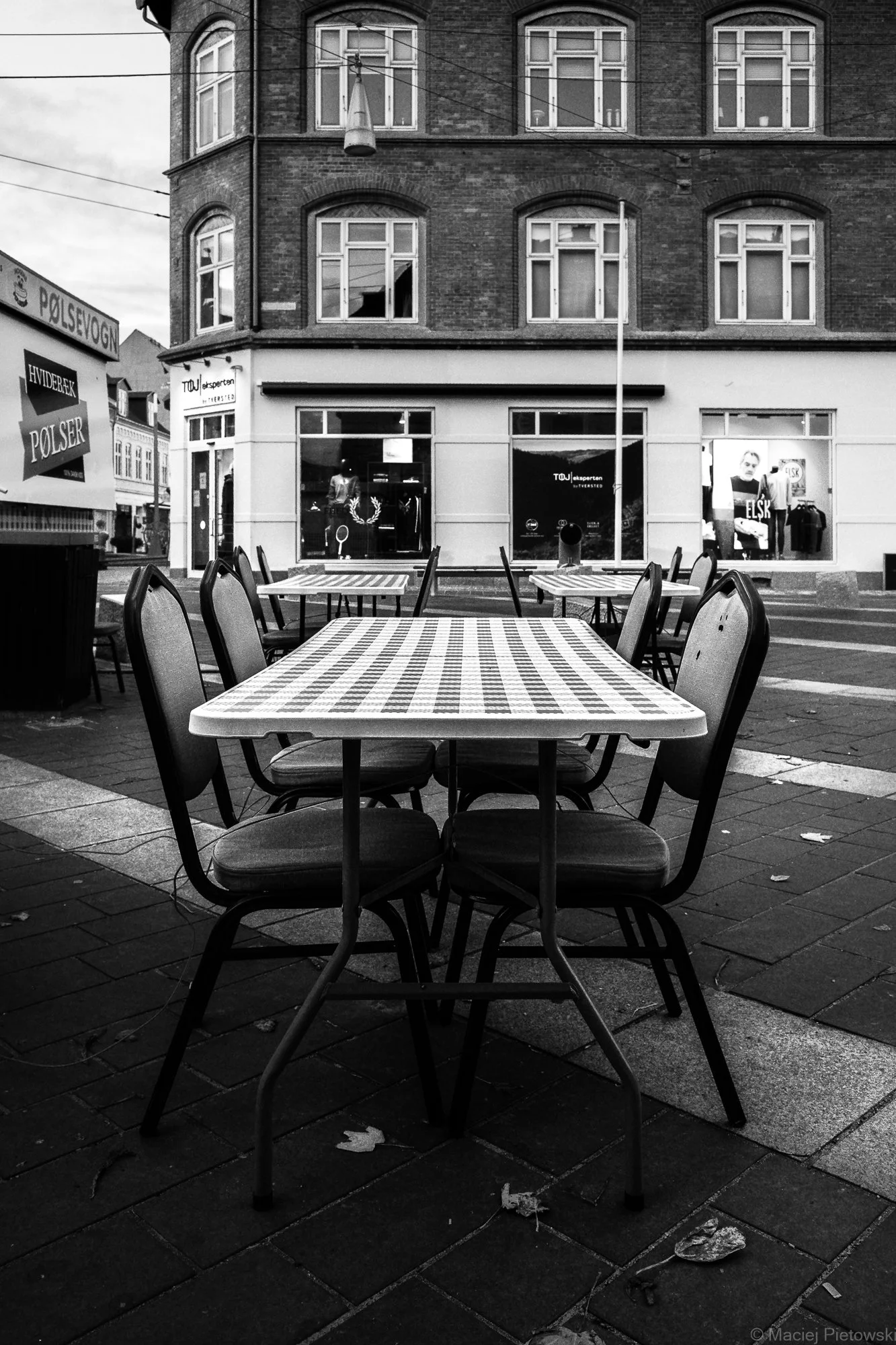Empty outdoor cafe with checkered tablecloths and chairs on a sidewalk, in front of storefronts and a brick building.