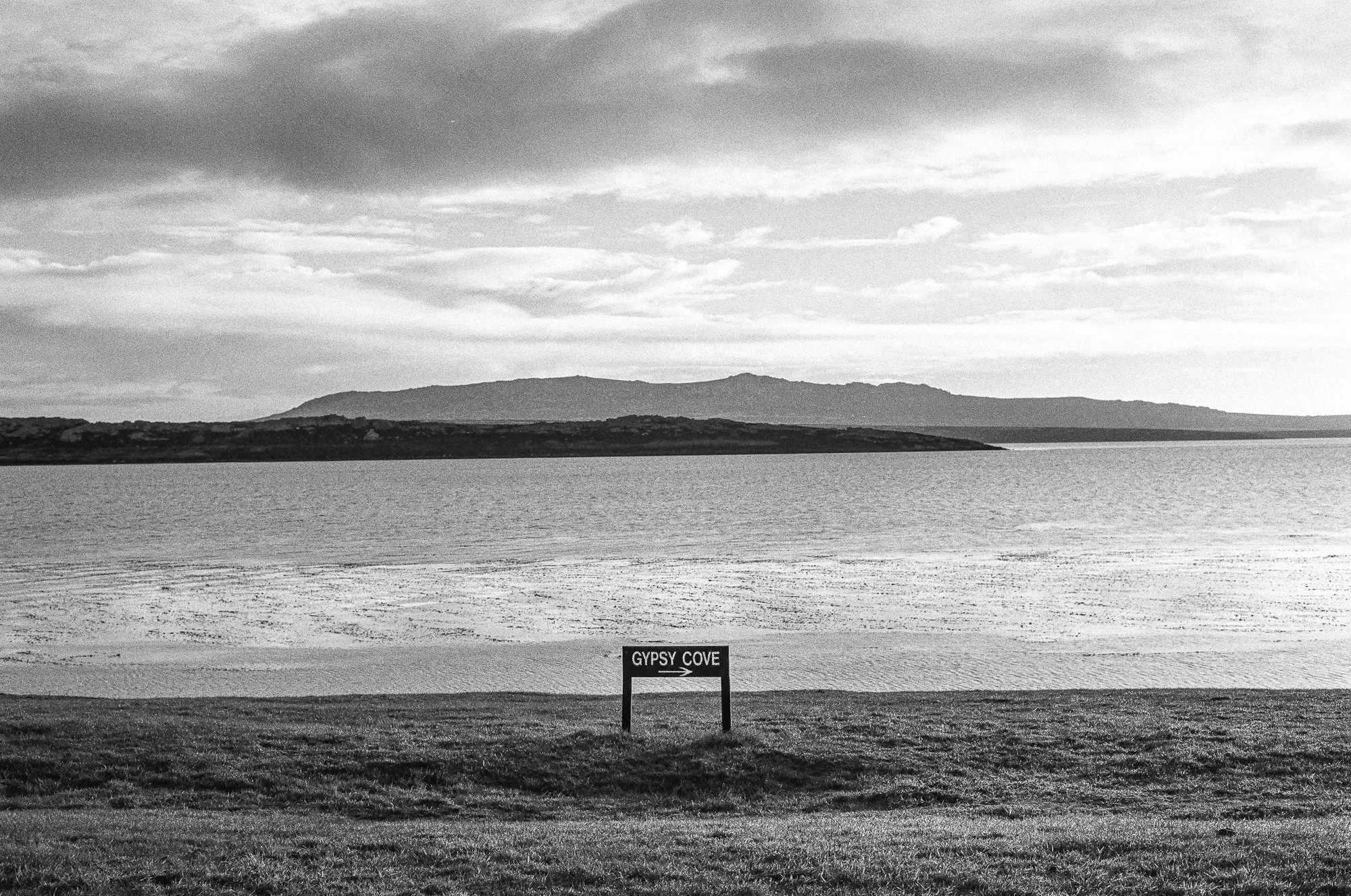 A black and white photograph of a large body of water with a distant shoreline and mountains in the background. There is a sign in the foreground that reads 'GYPSY COVE' with an arrow pointing to the right.