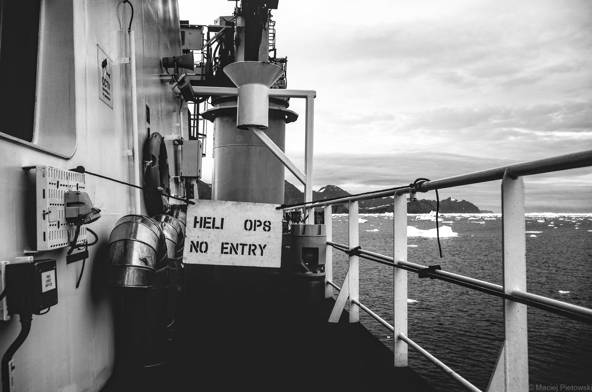 Photo of a ship's deck with equipment and safety signage, overlooking an ocean with ice and mountains in the distance, in black and white.
