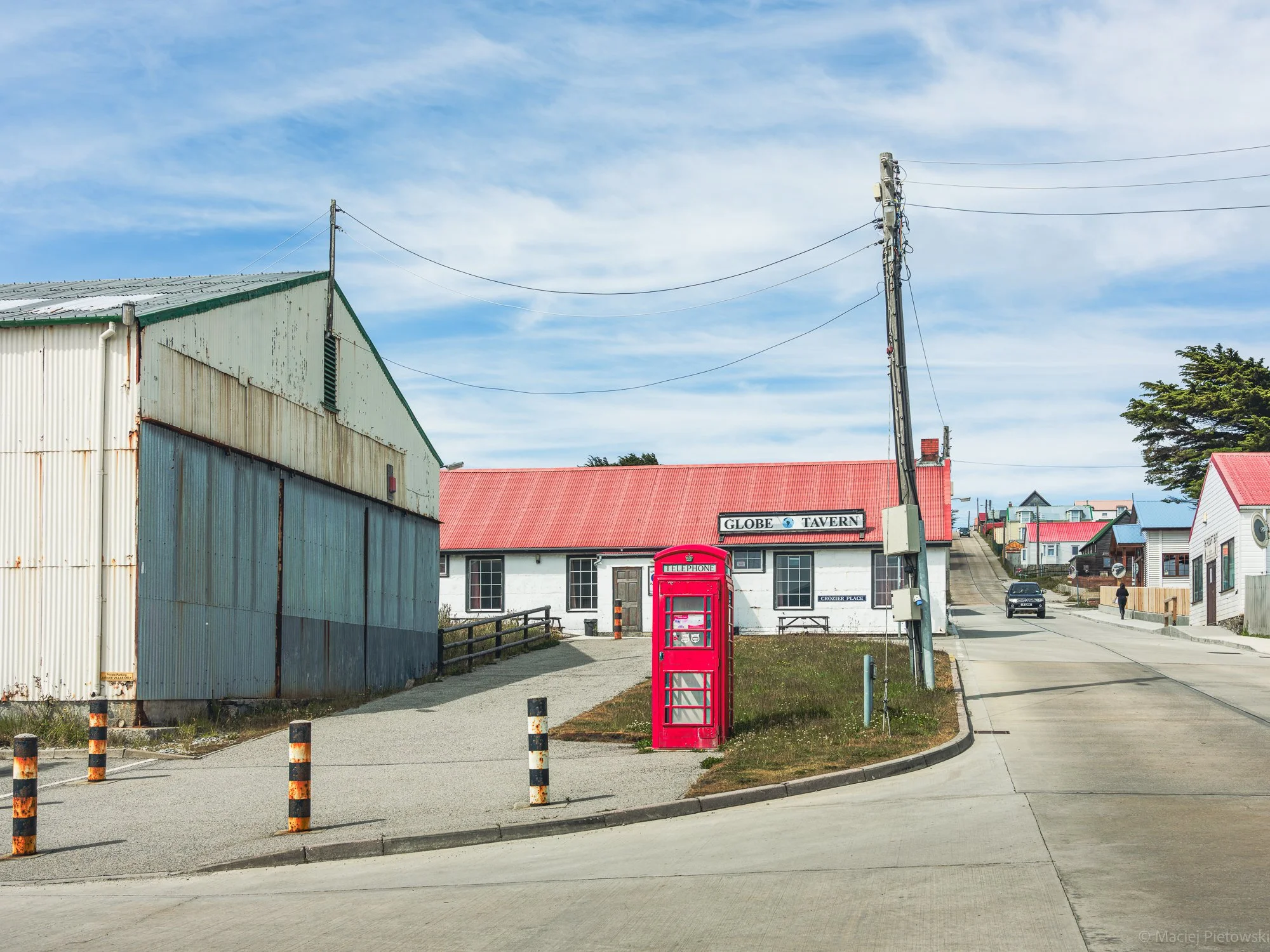 A small town street with a red telephone booth in the foreground and buildings with colorful roofs in the background. There is a power pole with wires and a sign that reads 'GLOBE TAVERN'. The sky is partly cloudy with light blue and white clouds.