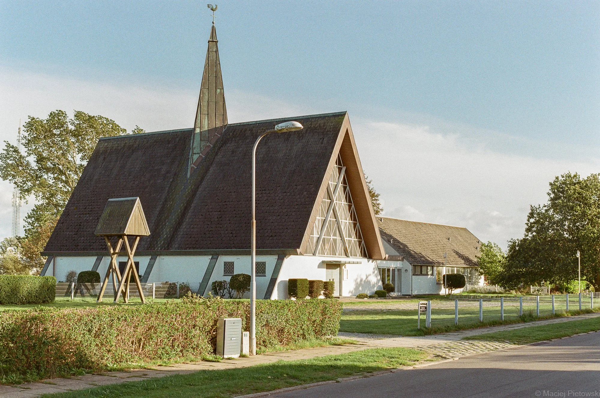 A church with a steep, black, shingled roof and a tall, pointed spire topped with a weather vane. There is a streetlamp near the church, and a small wooden structure on stilts. The church has large triangular windows and is surrounded by well-maintai