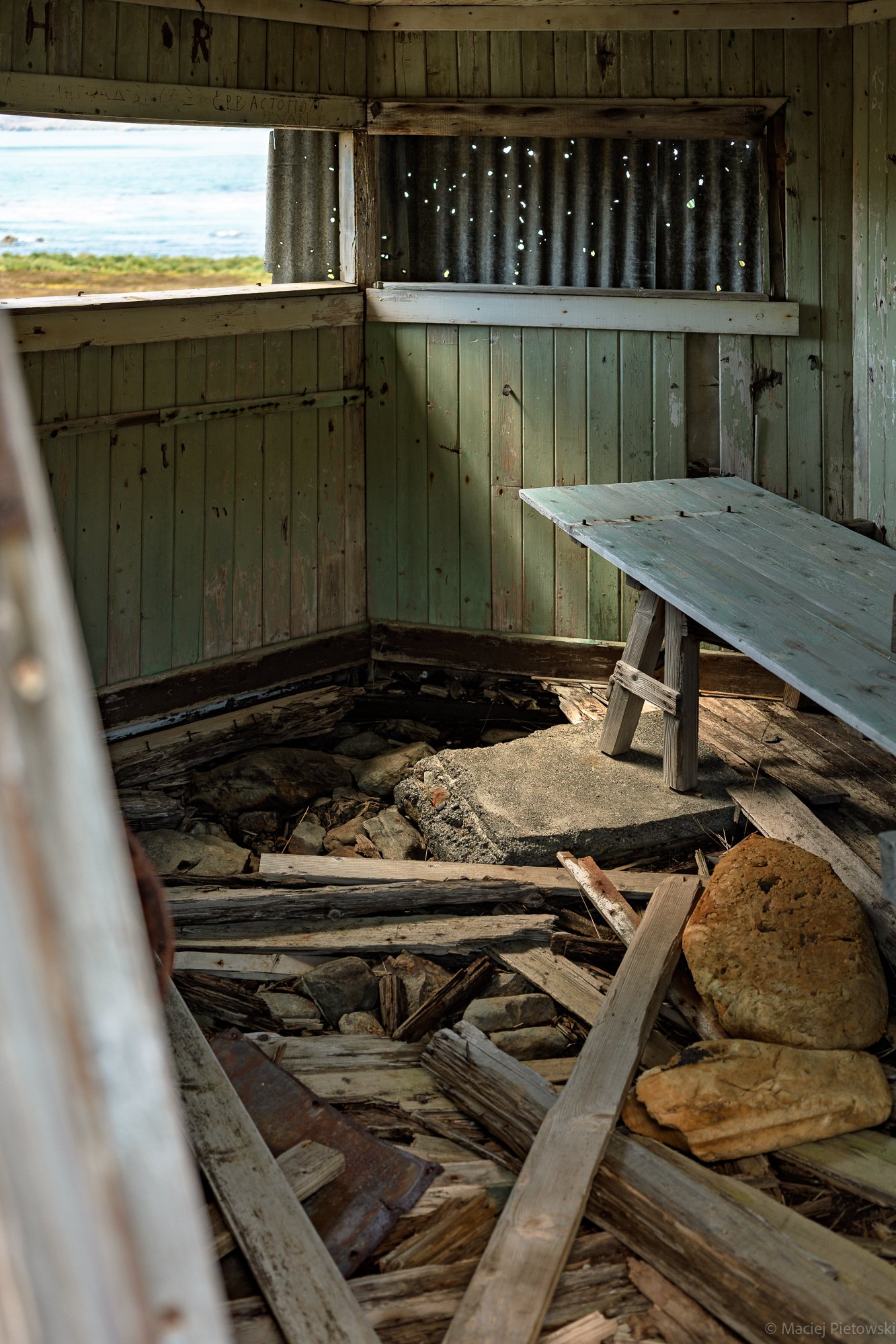 Inside an abandoned wooden structure, possibly a lookout or beach shelter, with weathered green planks, a wooden bench, and a hole in the floor revealing rocks below. Through the window, a view of the beach, ocean, and sky is visible, with corrugated