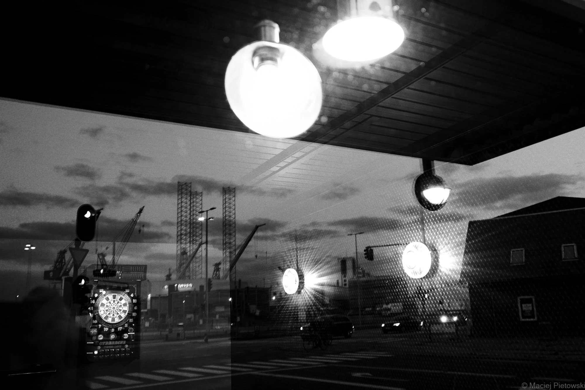Outdoor scene at dusk or night with illuminated streetlights and digital dartboard outside, reflected on glass window with a patterned surface.