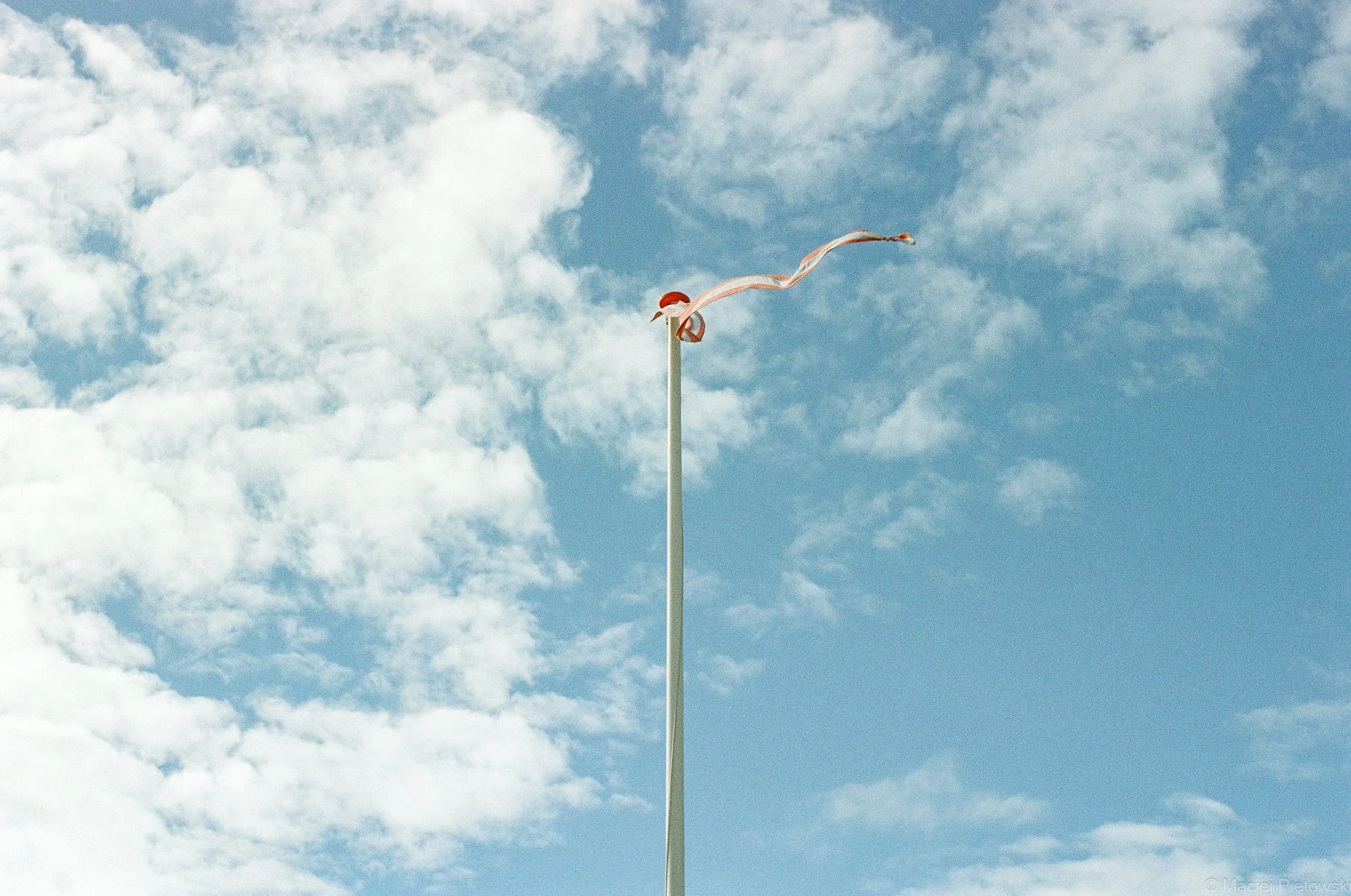 A tall flagpole with a weather vane at the top, blowing in the wind, against a partly cloudy sky.