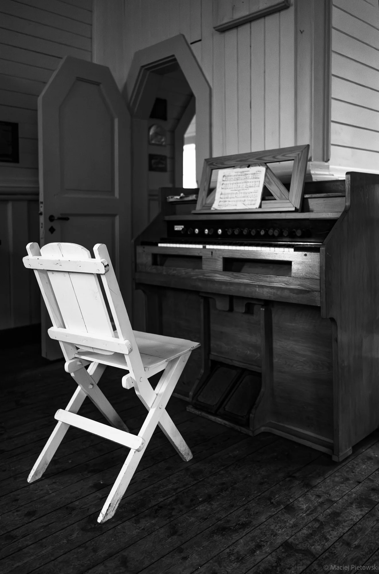 Black and white photo of a wooden church interior with a white folding chair in the foreground, a wooden cabinet with an open lid in the background, and a piano with sheet music on it.