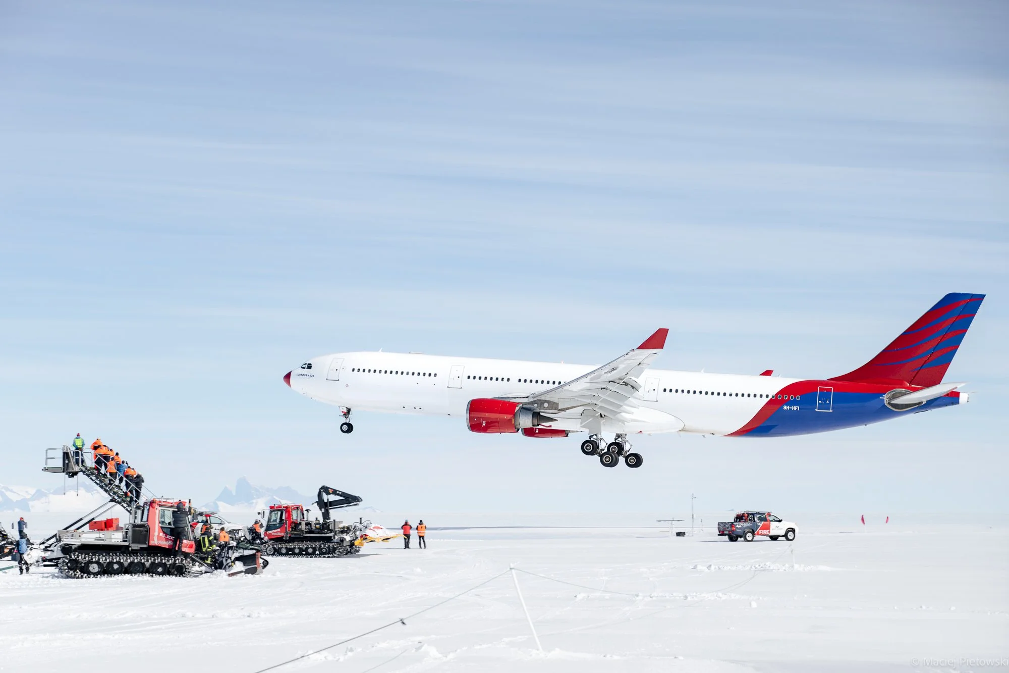 An airplane is in mid-air during takeoff or landing at an airport in a snowy, cold environment. Several ground crew members and vehicles are present below, preparing or assisting with the aircraft operation.