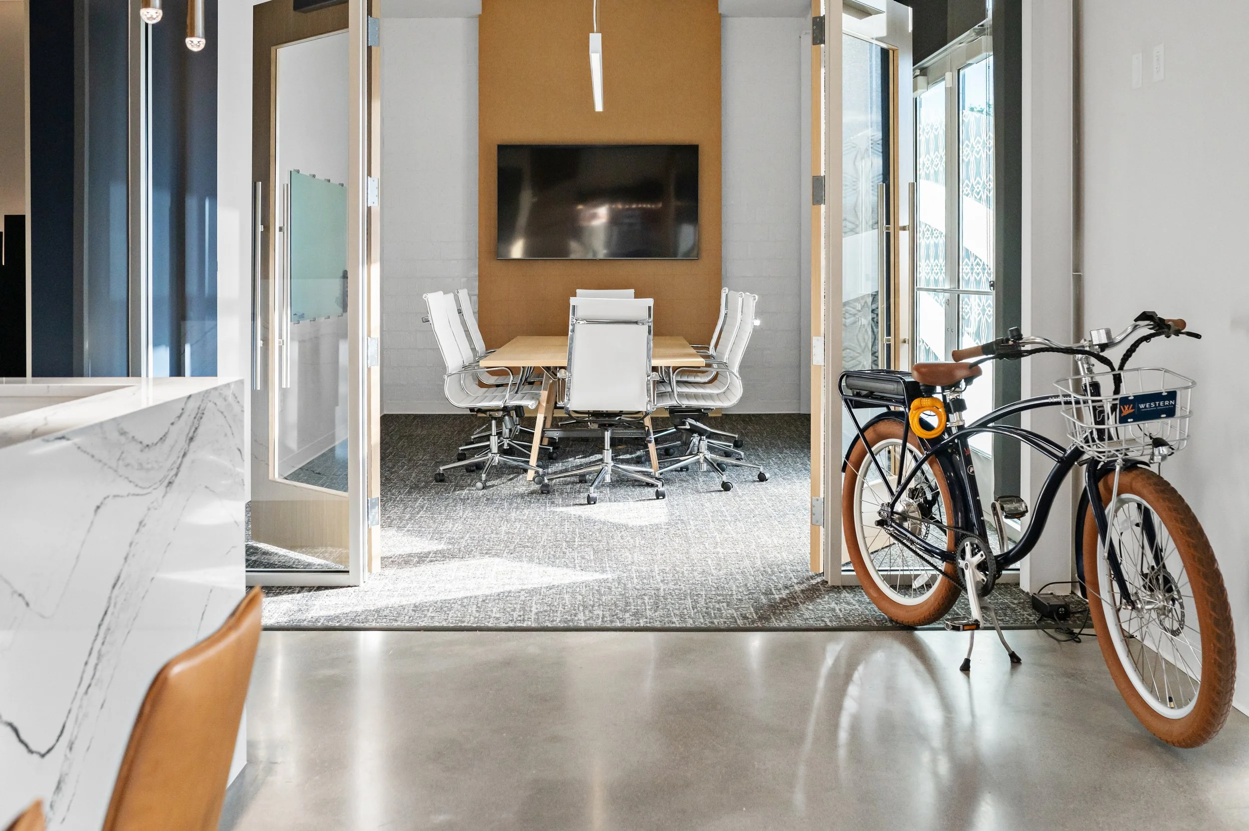 Western Insurance view through a doorway into a conference room with a large television mounted on a brown wall, a wooden table, white chairs, and two bicycles parked inside on the right side.