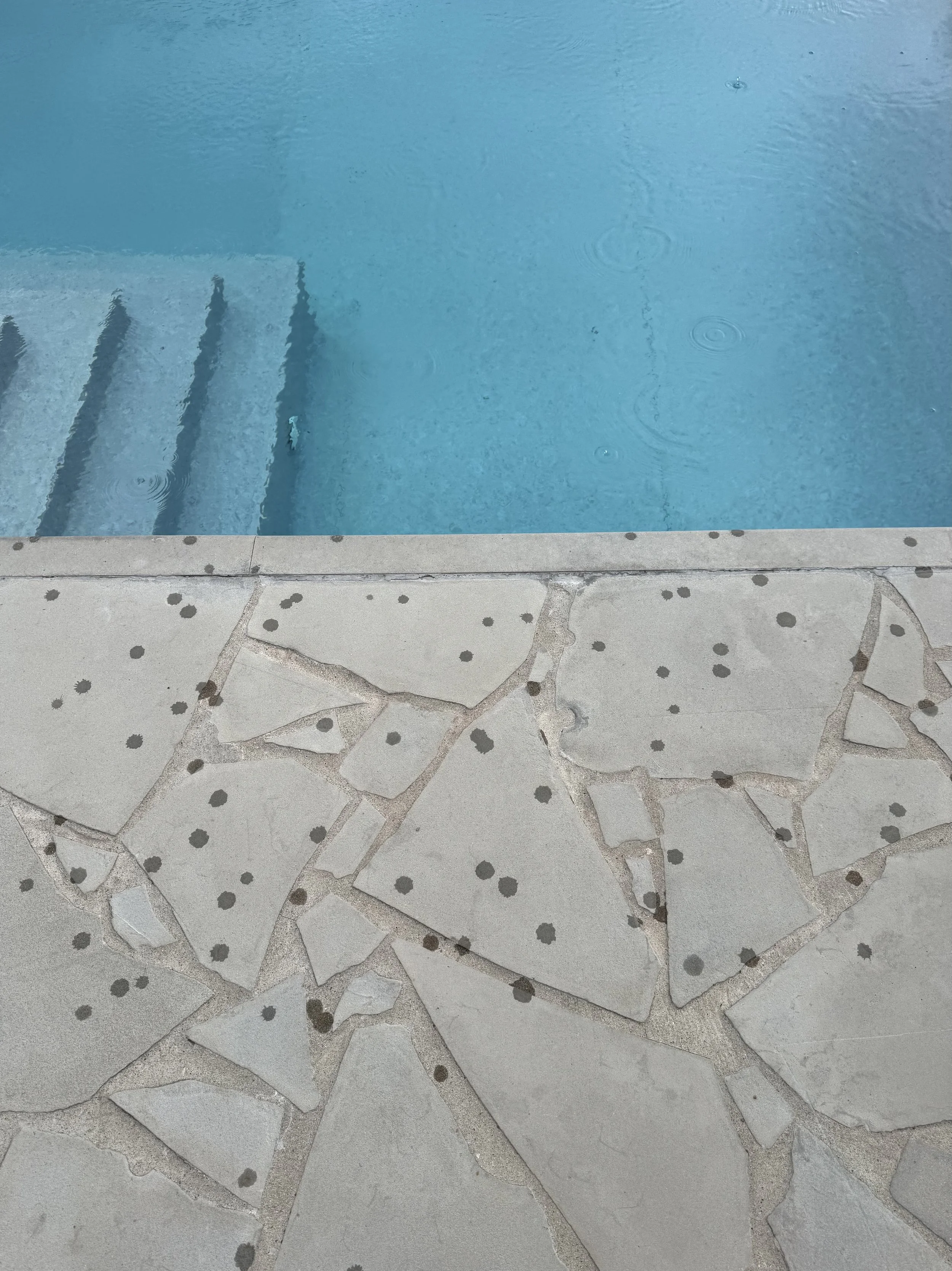 Minimalist view of the pool at Sesta Terra Resort in Italy as rain ripples the water surface and dots the stones. Atmospheric landscape evokes deep calm and restorative wellness, reflecting a restrained modern edit.