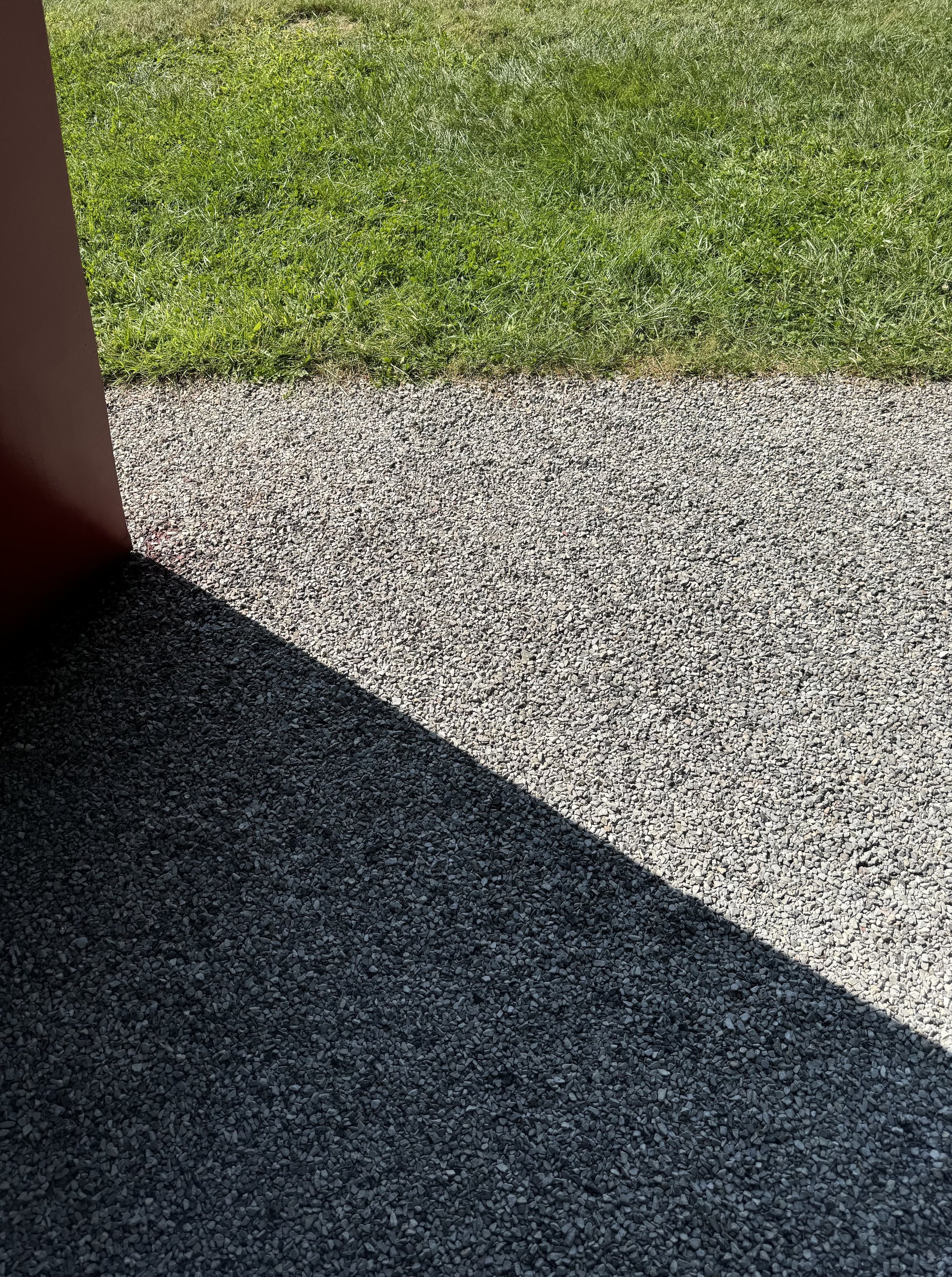 A graphic, minimalist architectural detail photograph at Philip Johnson's iconic Glass House. The composition captures a sharp geometric shadow cast across a pristine gravel pathway at its intersection with lush green lawn, a restrained modern edit.