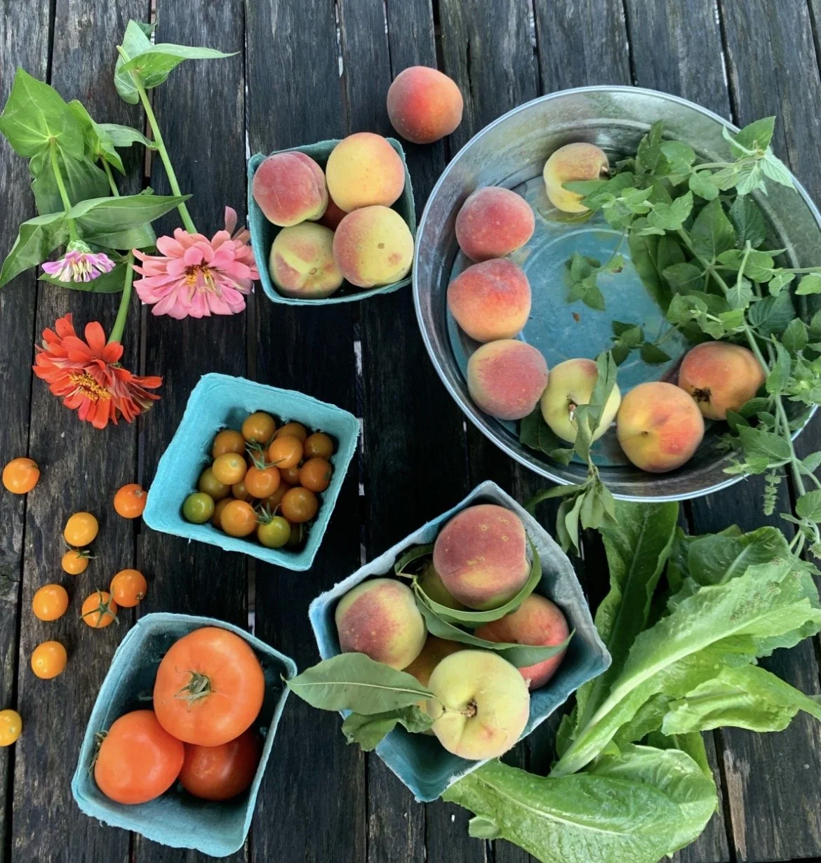 Assorted fresh fruits including peaches, tomatoes, and cherry tomatoes with some flowers and green leaves on a wooden table.