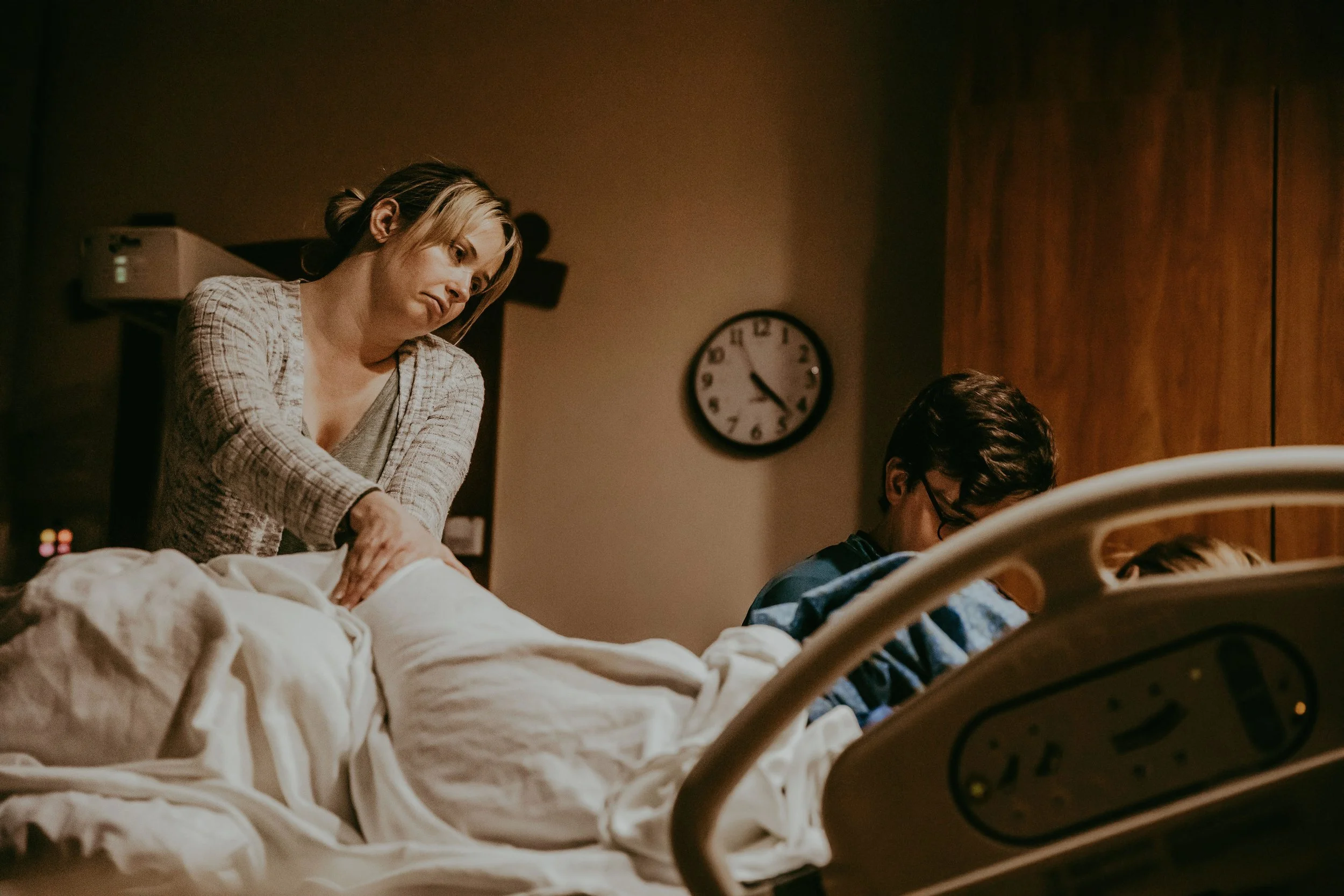 A woman in hospital gown leaning over a patient in a hospital bed, with a clock on the wall showing approximately 4:10.