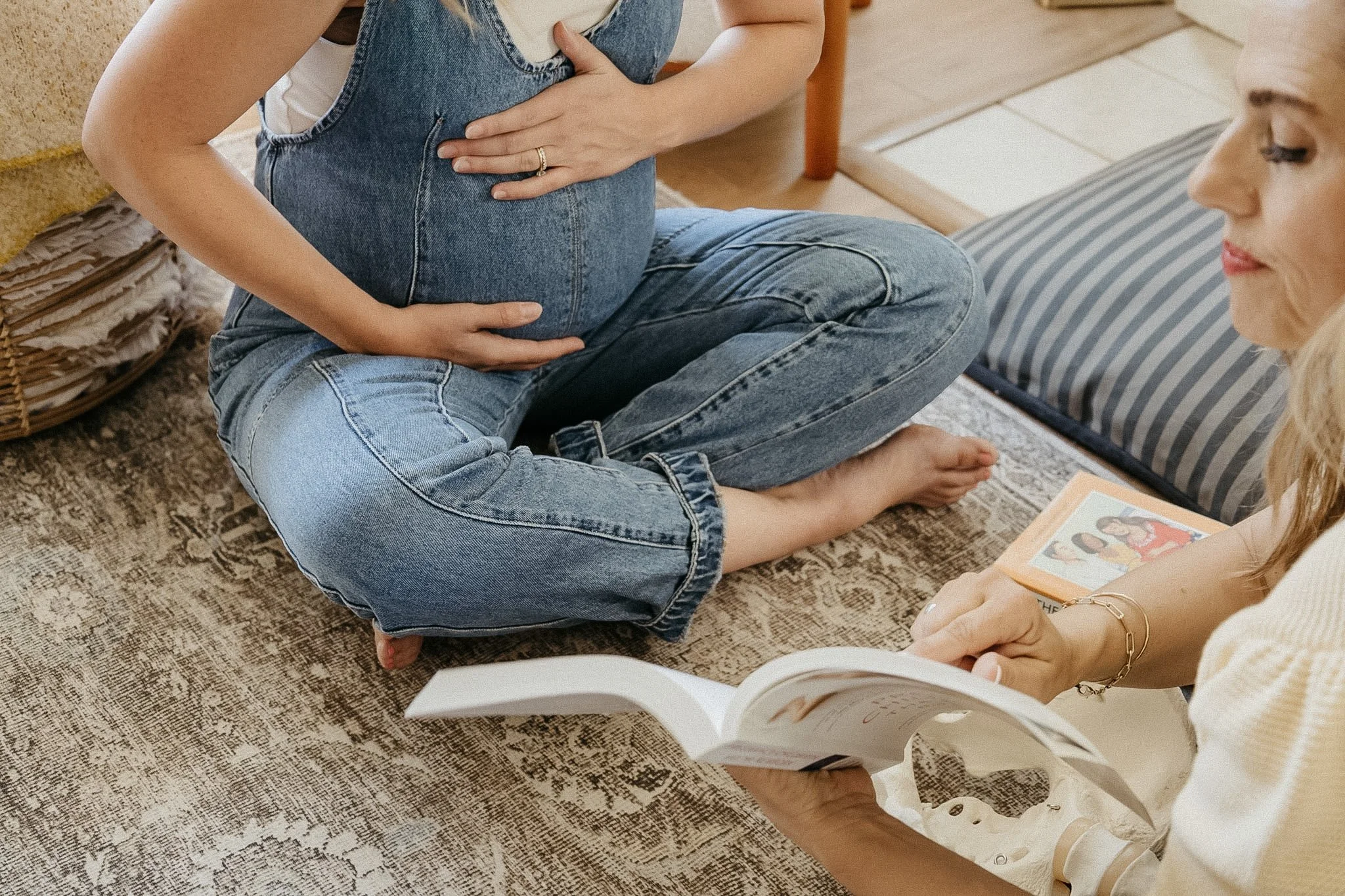 A pregnant woman sitting cross-legged on a rug, with her hand on her belly, and another woman nearby holding an open photo album.