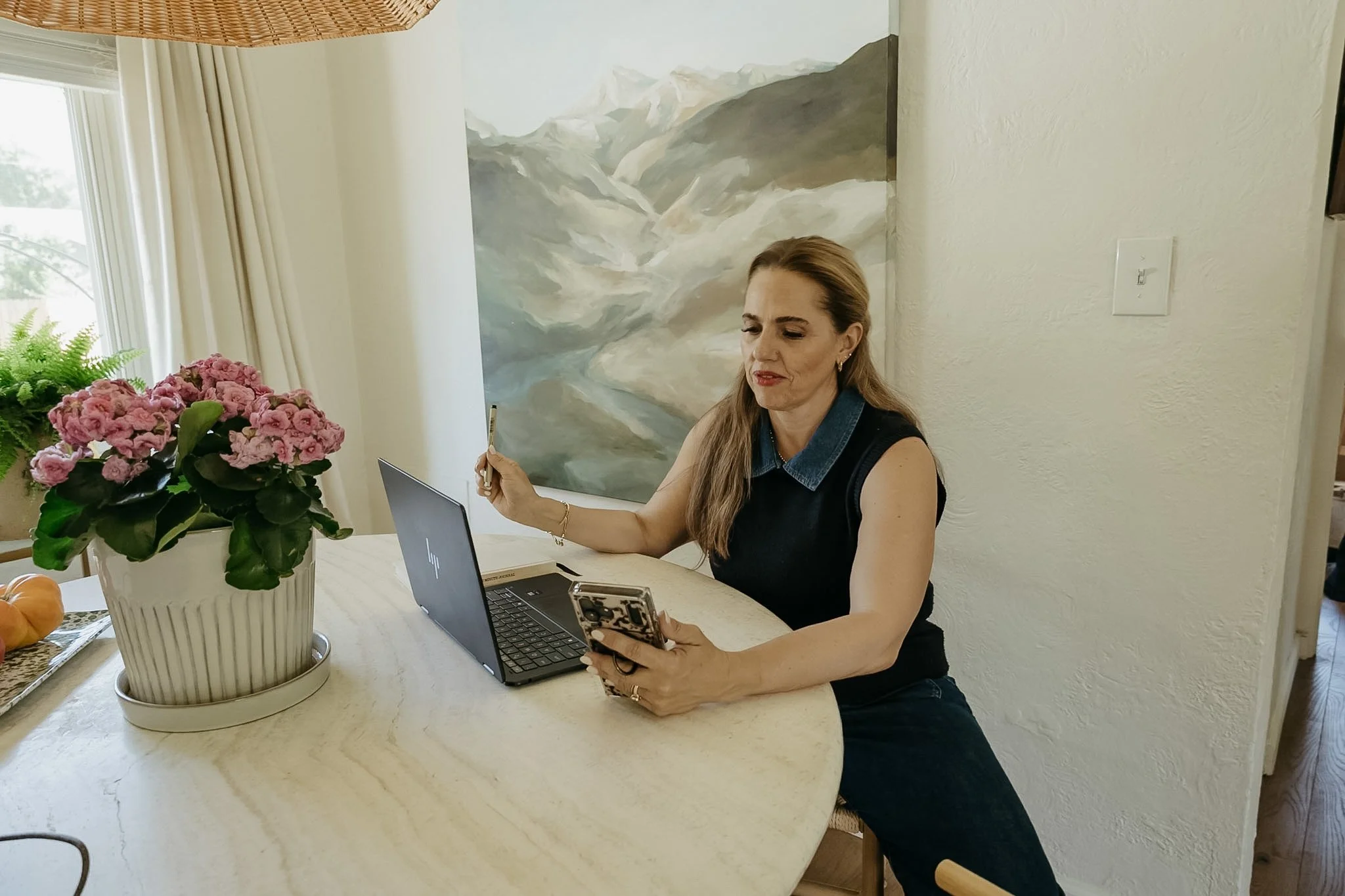 A woman with long blonde hair, wearing a sleeveless black top, sitting at a kitchen table. She is holding a smartphone in her right hand and a pen in her left hand. There is a laptop in front of her and a large pink flowering plant on the table. A painting of mountains hangs on the wall behind her.