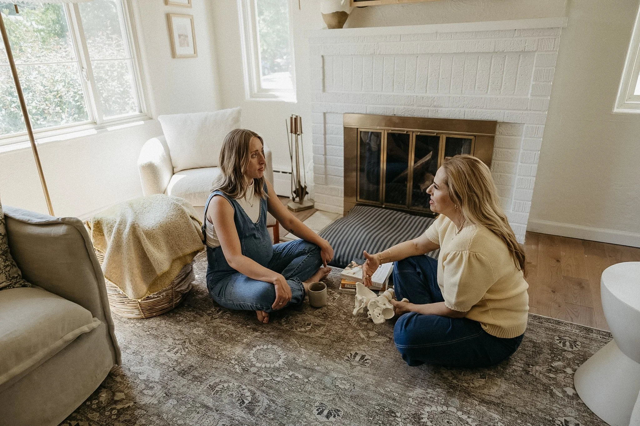 Two women sitting on the floor having a conversation in a living room near a fireplace, holding hands, with books, a skull model, and cups around them.
