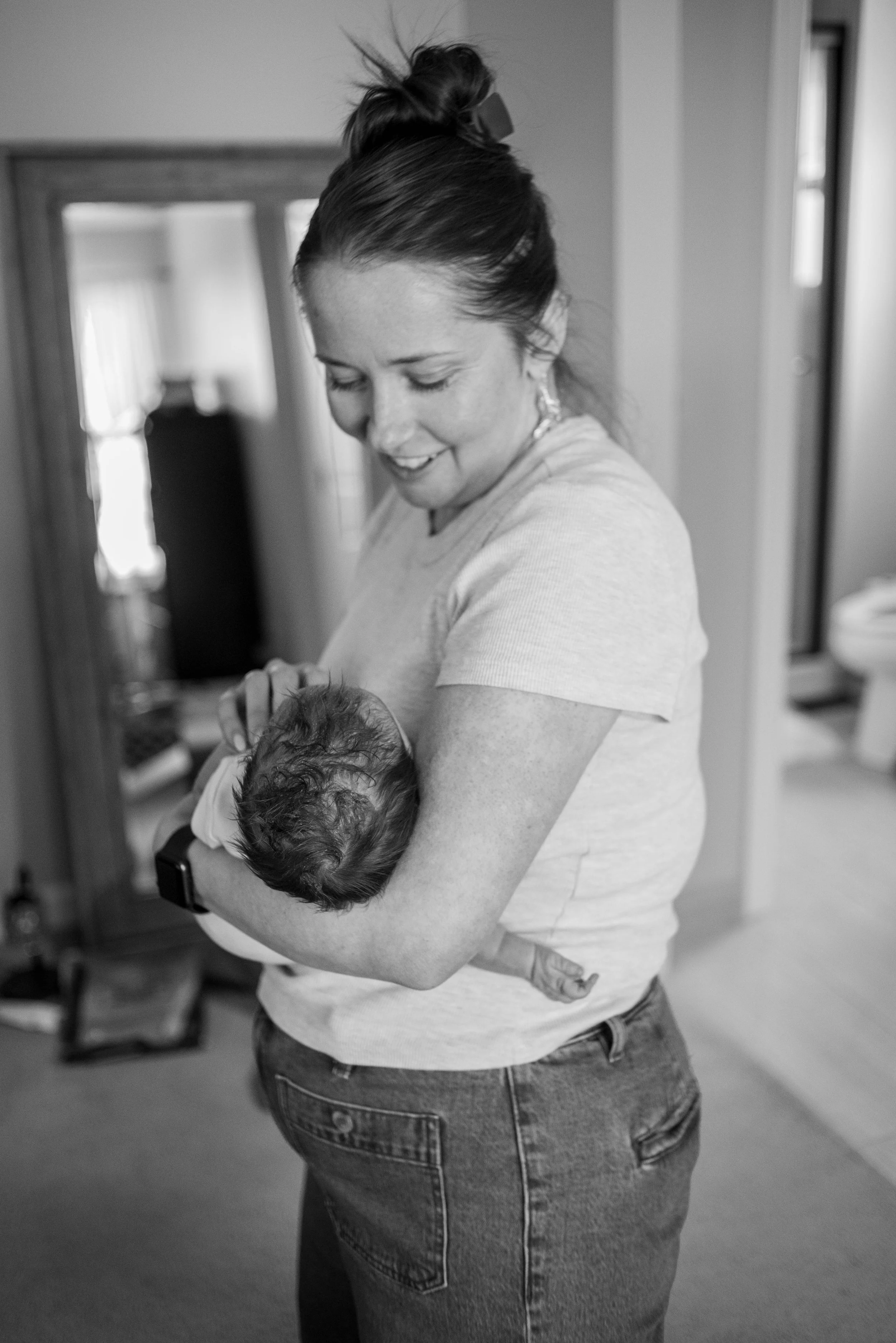 A woman with hair in a bun holding a sleeping baby in her arms, standing in a room with a mirror and furniture in the background.