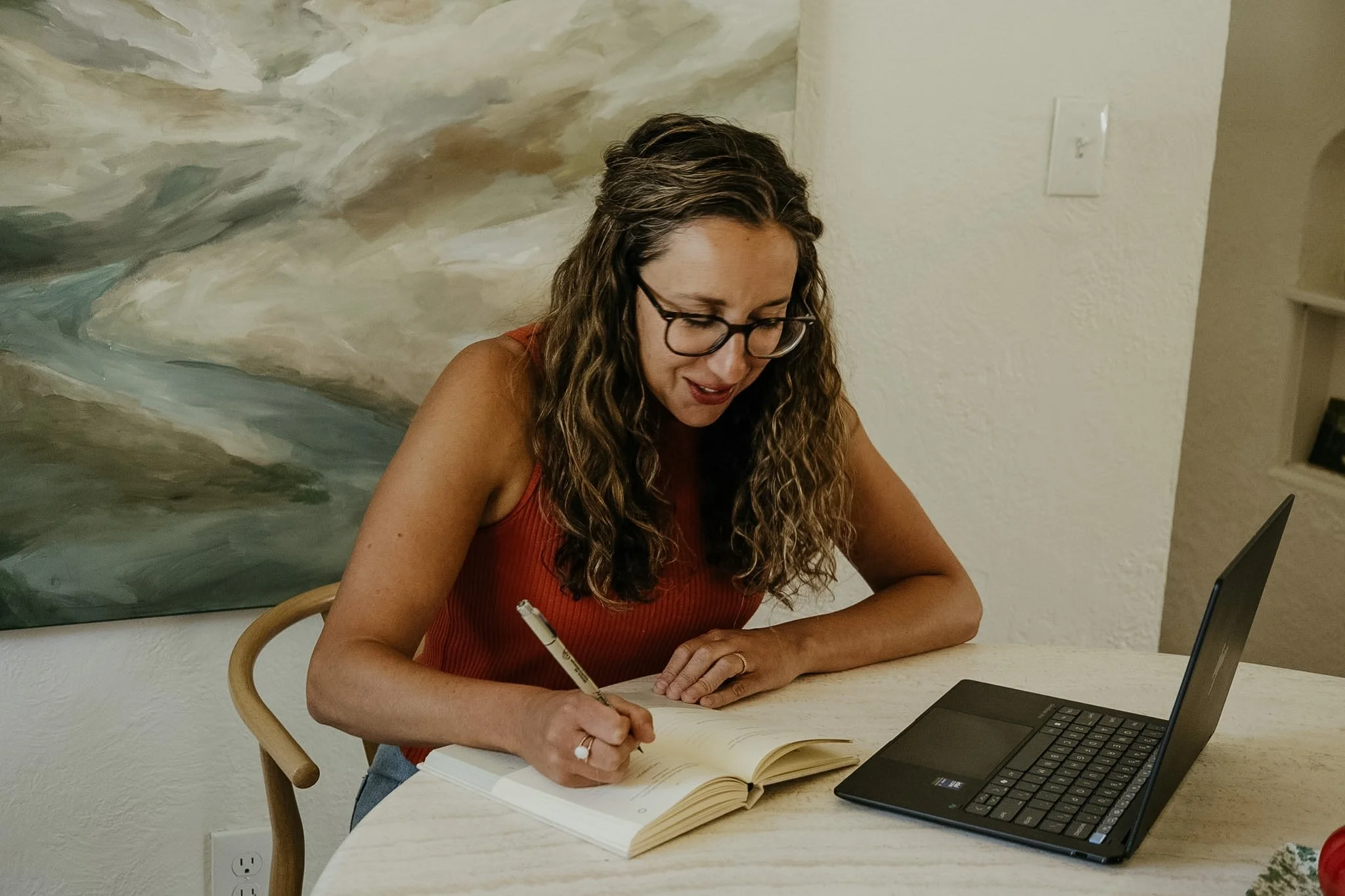 A woman with curly hair and glasses writing in a notebook at a table with a laptop nearby, sitting in front of a large abstract painting on the wall.