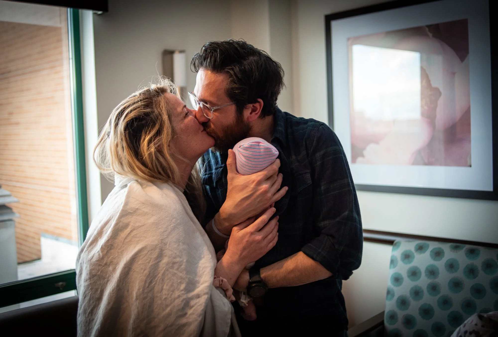 A couple sharing a kiss while holding a newborn baby. The scene is indoors, with a window and artwork in the background.