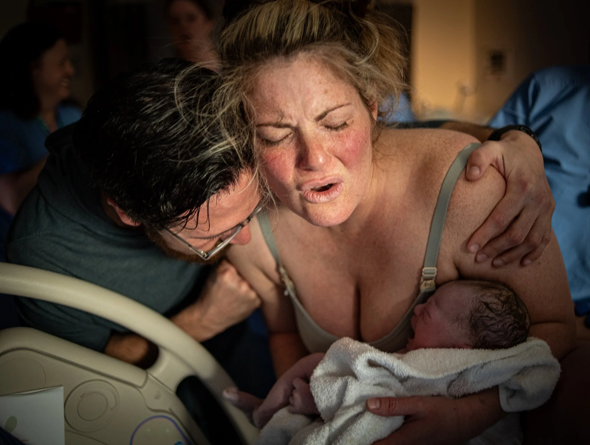 A woman with blonde hair, eyes closed, looks emotional while hugging a person during childbirth. The person being embraced has dark, wet hair and a head covered with a towel, likely a newborn. Medical staff and a baby blanket are visible in the background.