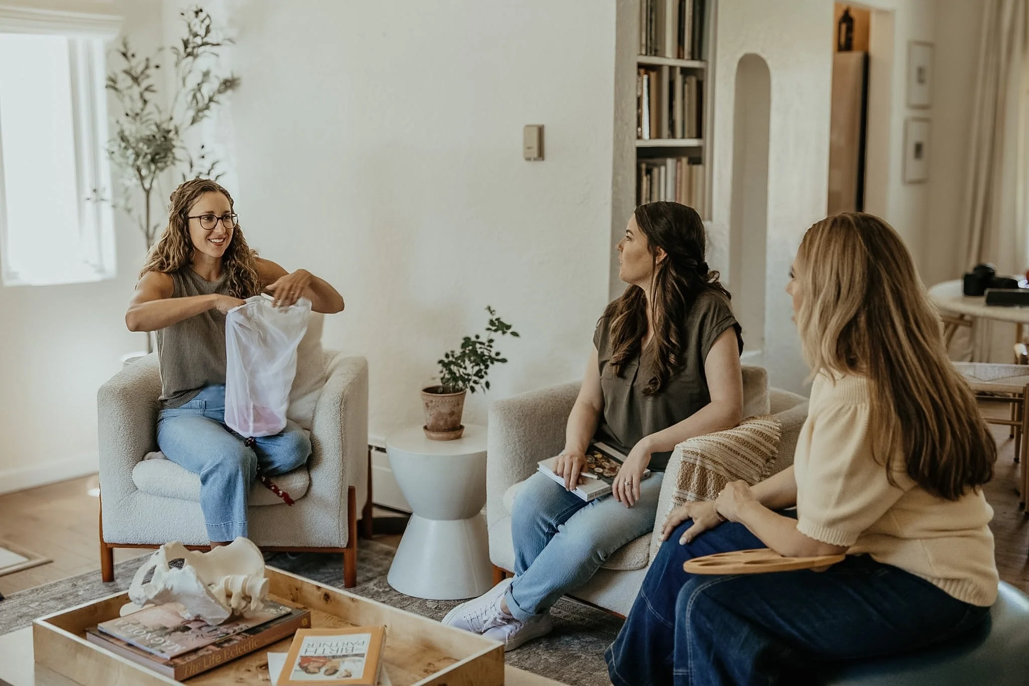 Four women sitting in a living room, one woman standing and smiling as she opens a gift bag. The other three women are seated on a couch and chair, observing and listening. The room is decorated with books, plants, and a coffee table with magazines and papers.