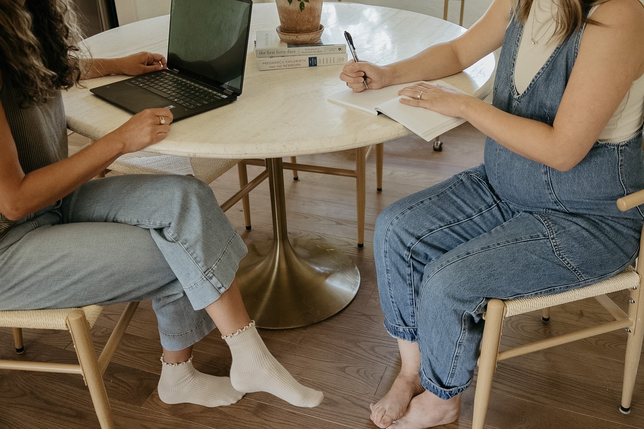 Two women sitting at a round table, one working on a laptop and the other writing in a notebook. Books and a potted plant are on the table.
