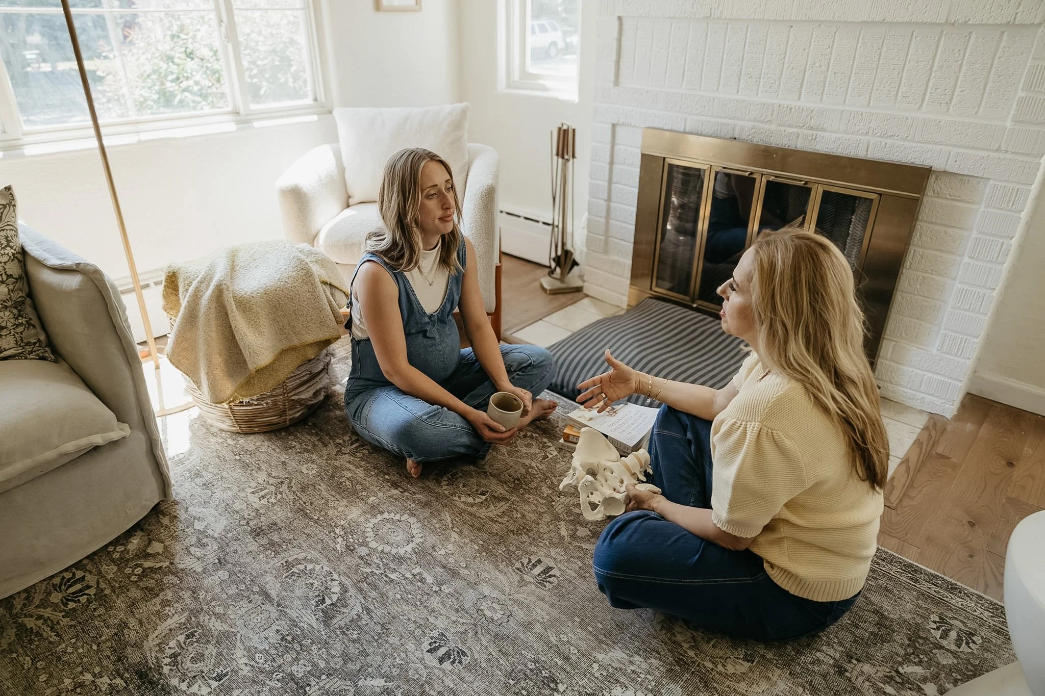 Two women sitting on the floor facing each other in a living room, engaged in conversation. One woman is holding a mug, and there are books and tissue papers nearby. The room has large windows, a white brick fireplace, and a patterned rug.