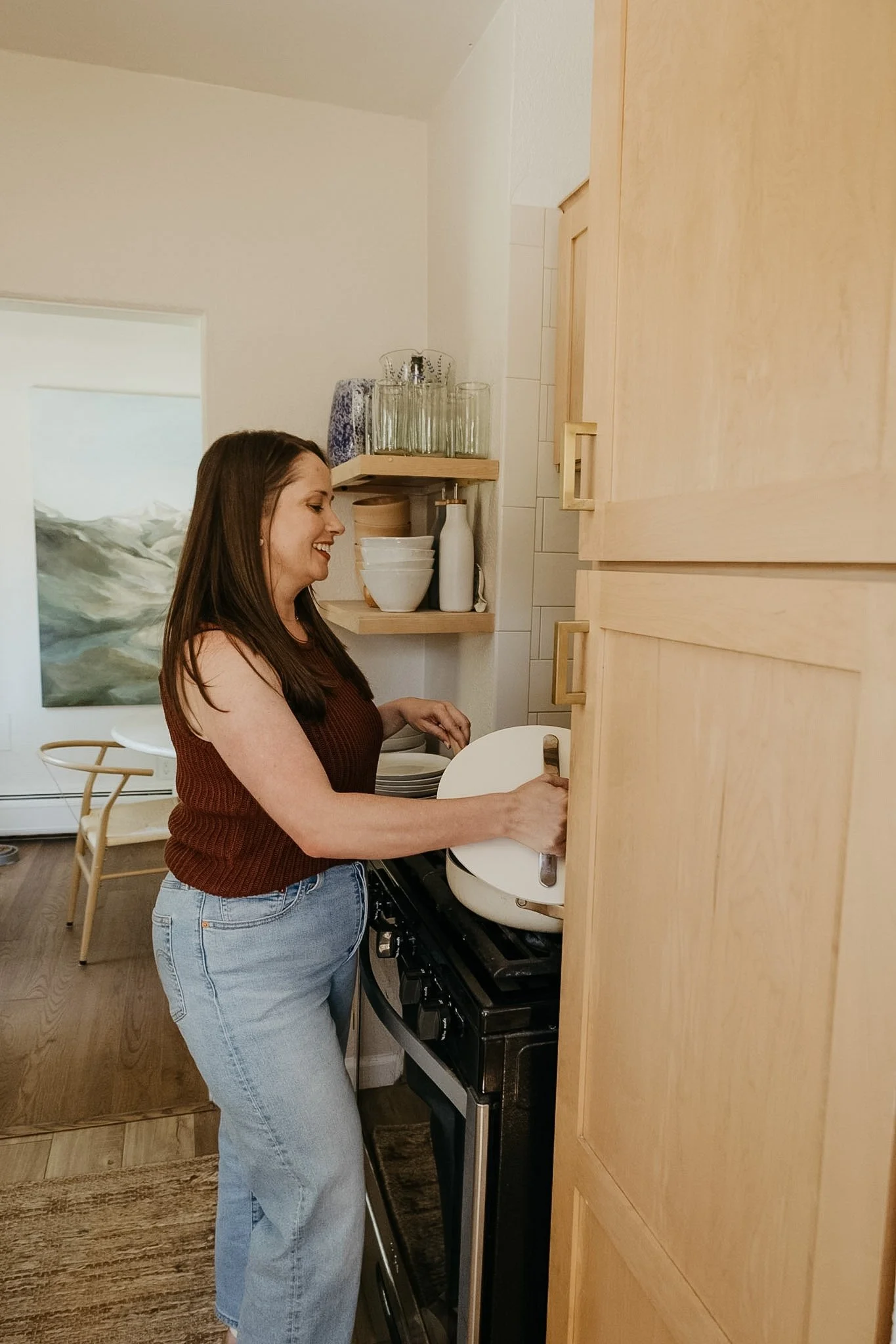 A woman with long brown hair wearing a sleeveless maroon top and light blue jeans is standing in a kitchen, using a stove. She is smiling and appears to be cooking or preparing food. The kitchen has light-colored wooden cabinets, a wooden shelf with glassware and dishes, and a painting hanging on the wall. The floor is wooden, and there is a chair with a woven back behind her.