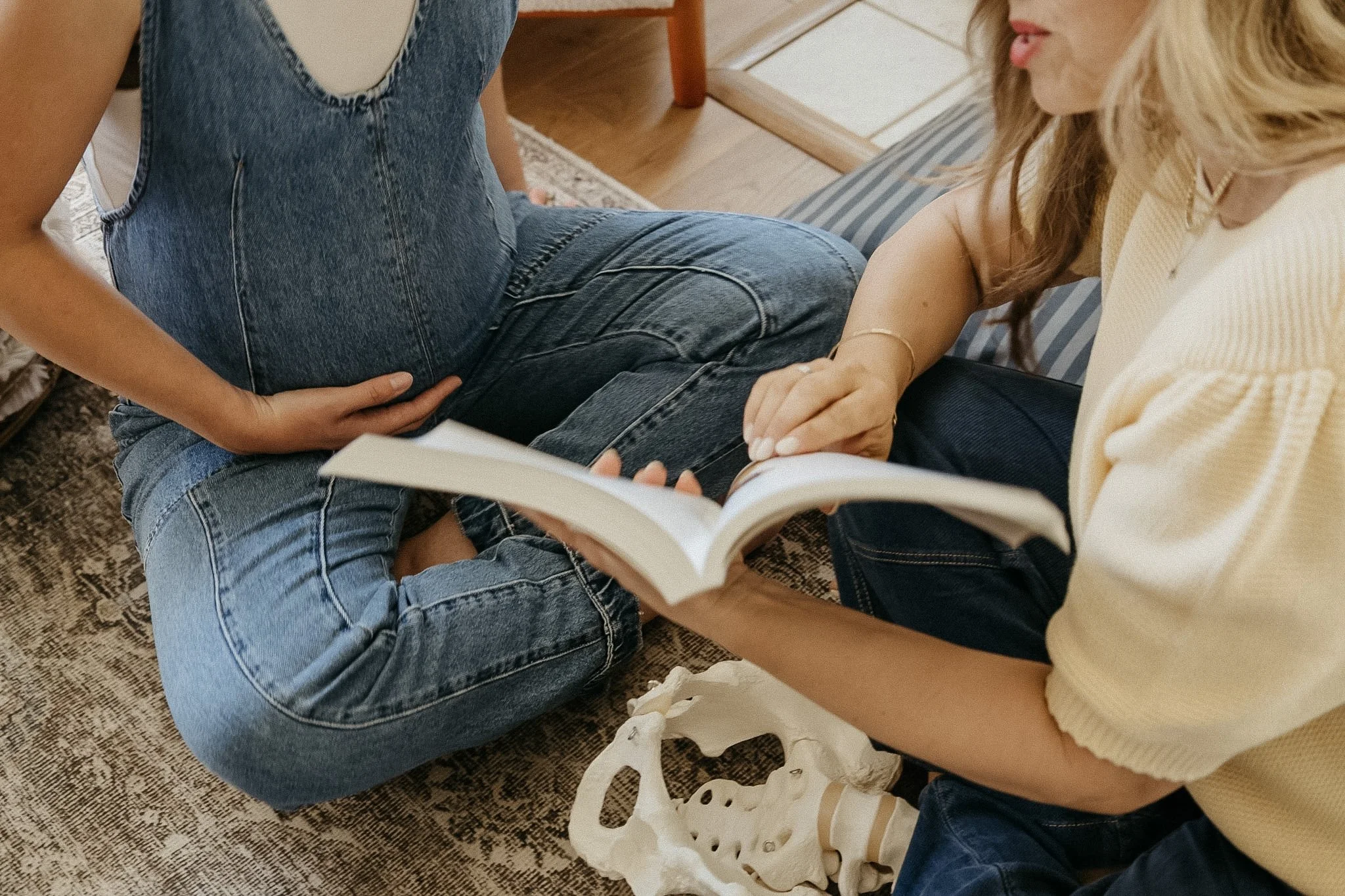 Two women sitting on the floor, one wearing a denim jumpsuit and the other a yellow sweater, examining a human skull and pelvic bone.