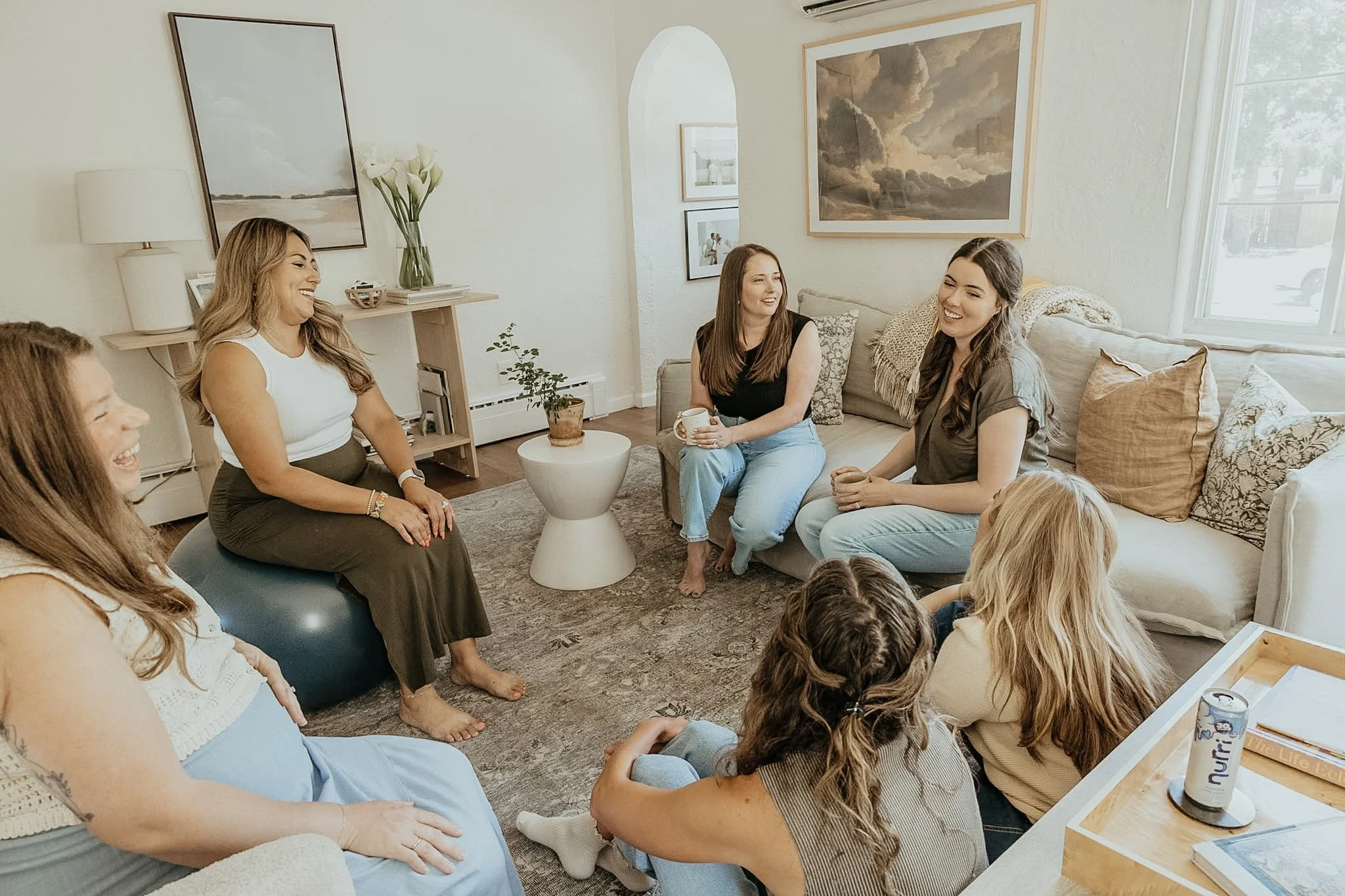 Group of women laughing and chatting in a cozy living room with neutral decor and large windows.